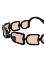 A close-up of ISSEY MIYAKE EYES UROKO sunglasses reveals distinctive interconnected black frames with pink-tinted lenses and a chain-like arm design, highlighting innovative Japanese craftsmanship.