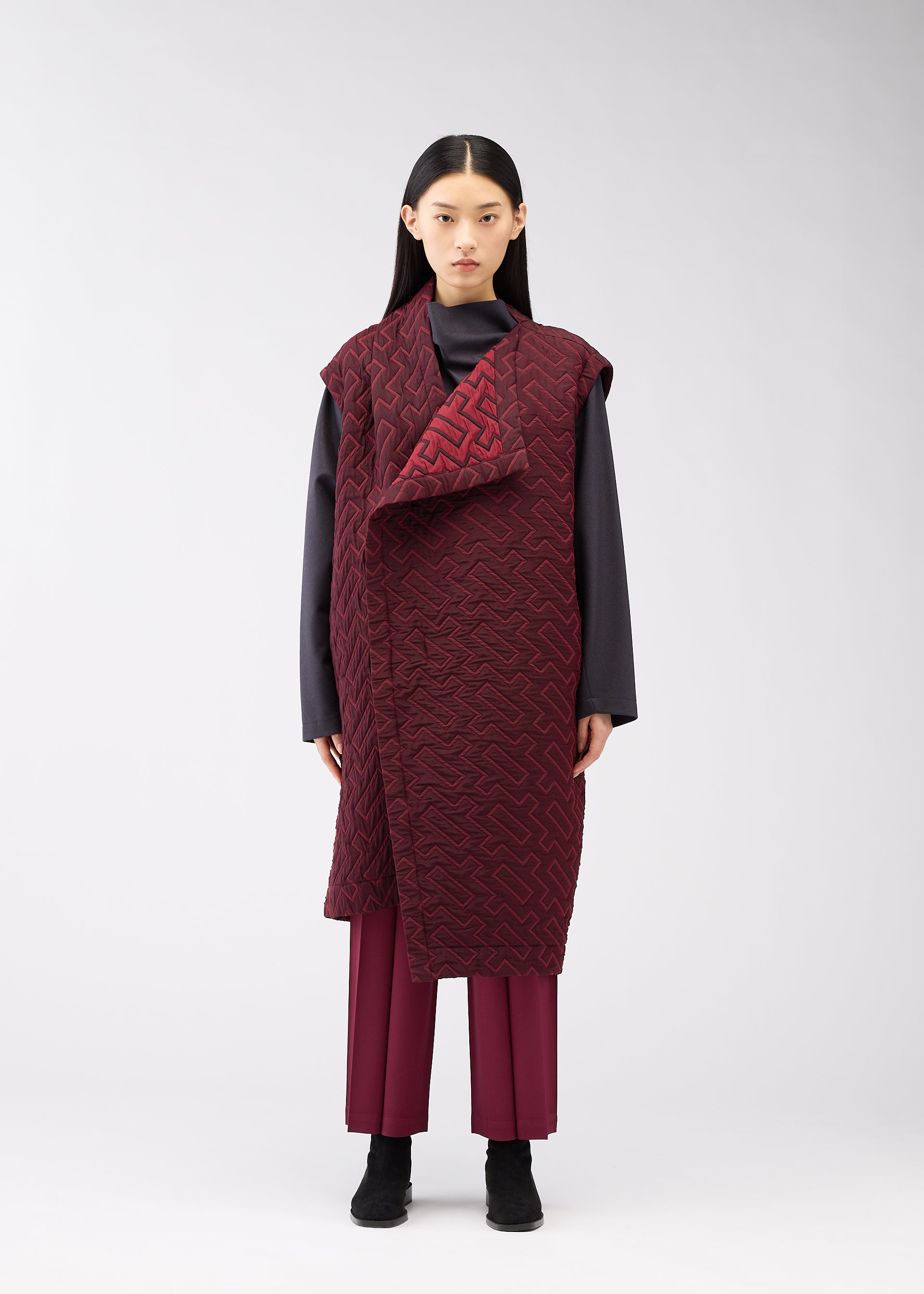 Zoomed view A woman with straight black hair and a neutral expression stands before a light gray backdrop, wearing the ONE CLOTH PUFF by ISSEY MIYAKE ONLINE STORE—a long burgundy jacquard vest over a dark gray top, matching pants, and black shoes.