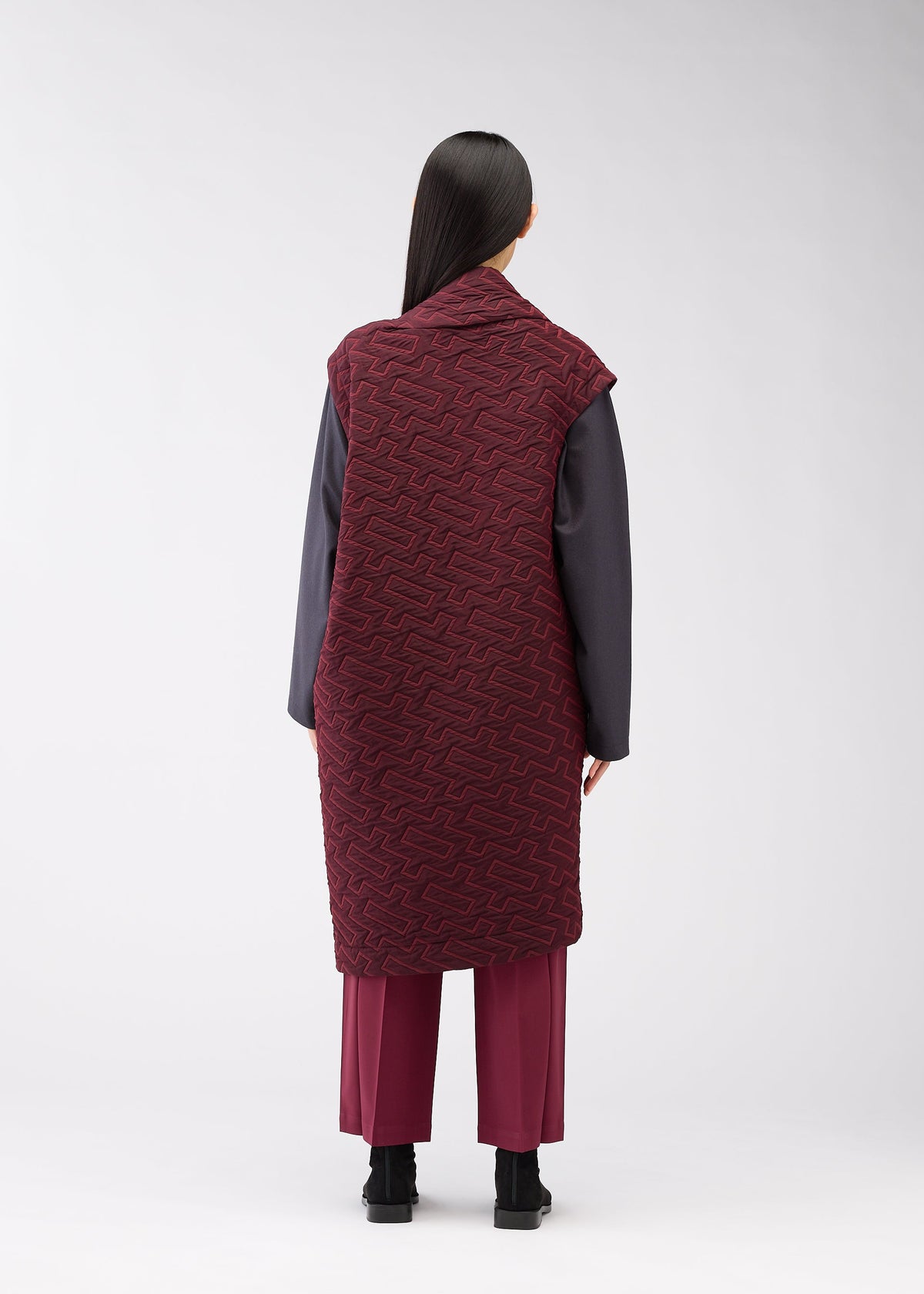 A person stands facing away, wearing the ONE CLOTH PUFF by ISSEY MIYAKE ONLINE STORE—a long maroon jacquard vest over a dark gray top and maroon pants. Their straight black hair and black shoes create a striking silhouette against a light background. view 6 of 8