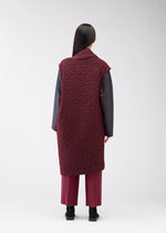 A person stands facing away, wearing the ONE CLOTH PUFF by ISSEY MIYAKE ONLINE STORE—a long maroon jacquard vest over a dark gray top and maroon pants. Their straight black hair and black shoes create a striking silhouette against a light background.