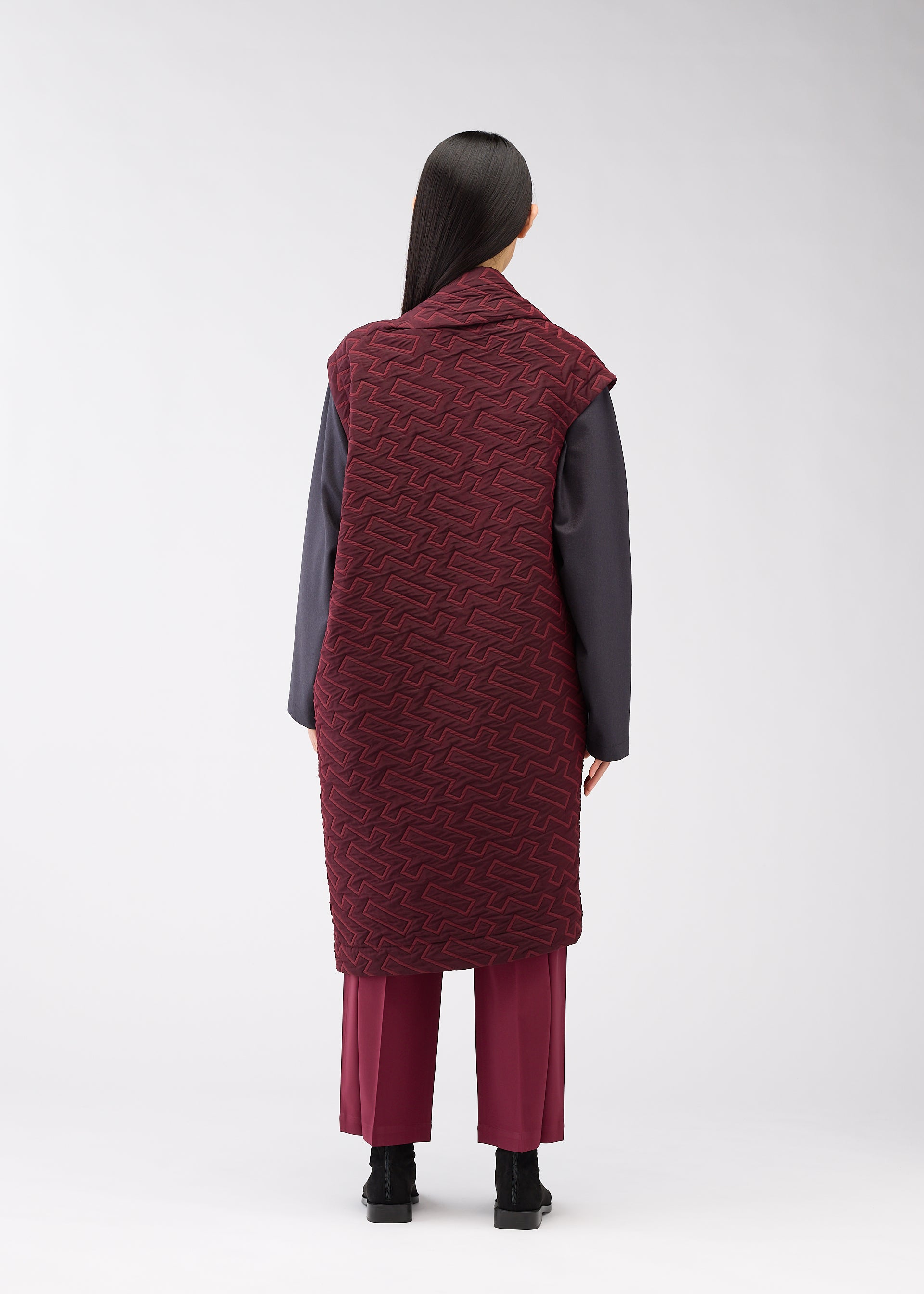 Zoomed view A person stands facing away, wearing the ONE CLOTH PUFF by ISSEY MIYAKE ONLINE STORE—a long maroon jacquard vest over a dark gray top and maroon pants. Their straight black hair and black shoes create a striking silhouette against a light background.