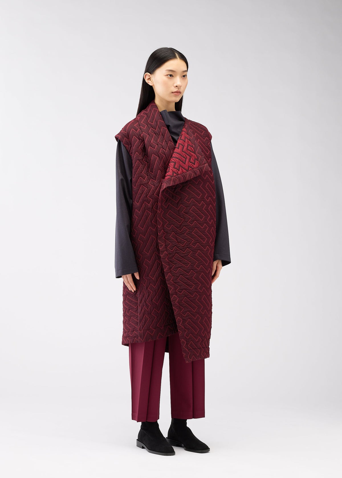 A woman with straight black hair models the ONE CLOTH PUFF by ISSEY MIYAKE ONLINE STORE— a long jacquard weave burgundy vest over a dark gray top and matching burgundy pants, set against a plain light gray background. view 7 of 8