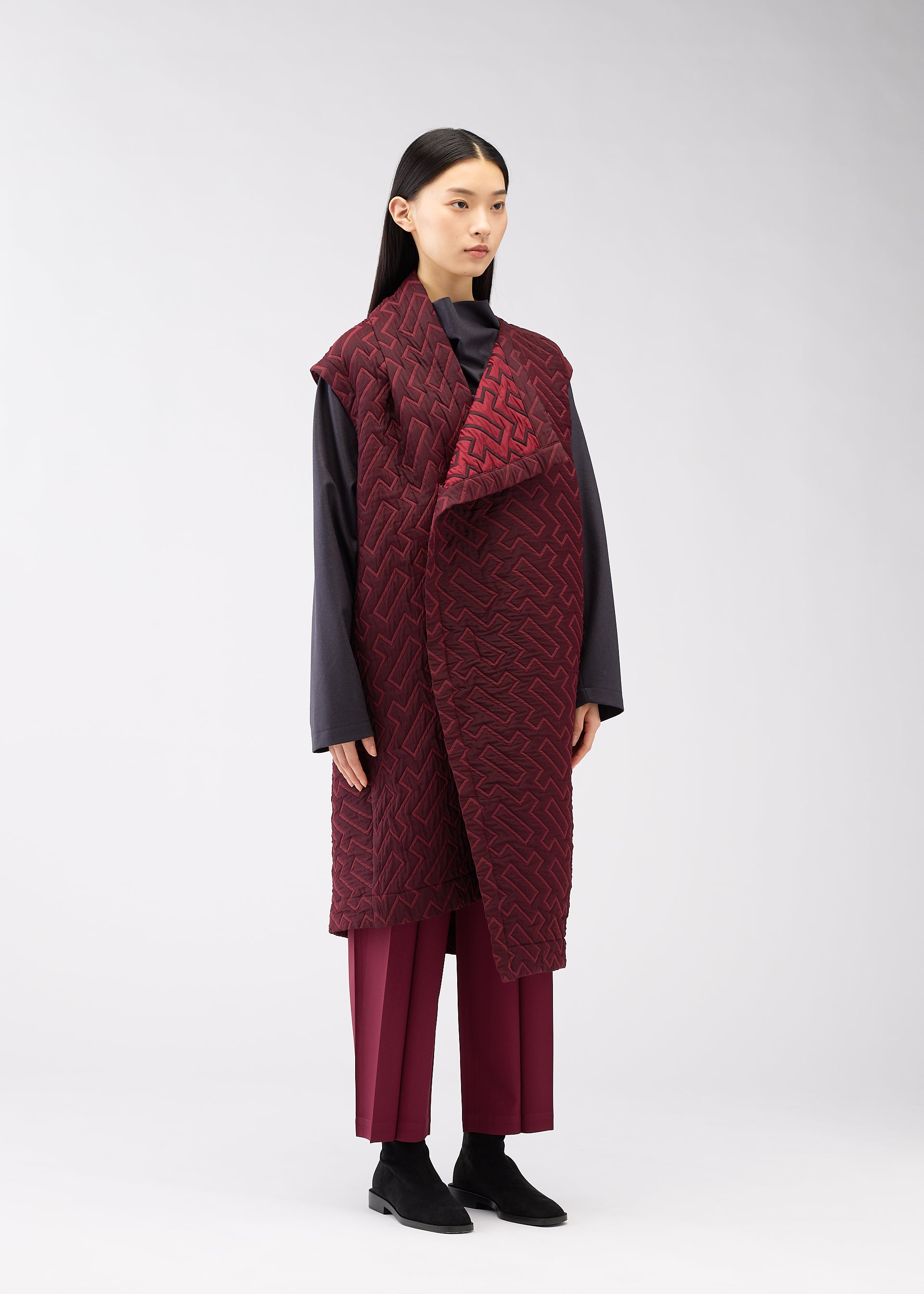 Zoomed view A woman with straight black hair models the ONE CLOTH PUFF by ISSEY MIYAKE ONLINE STORE— a long jacquard weave burgundy vest over a dark gray top and matching burgundy pants, set against a plain light gray background.