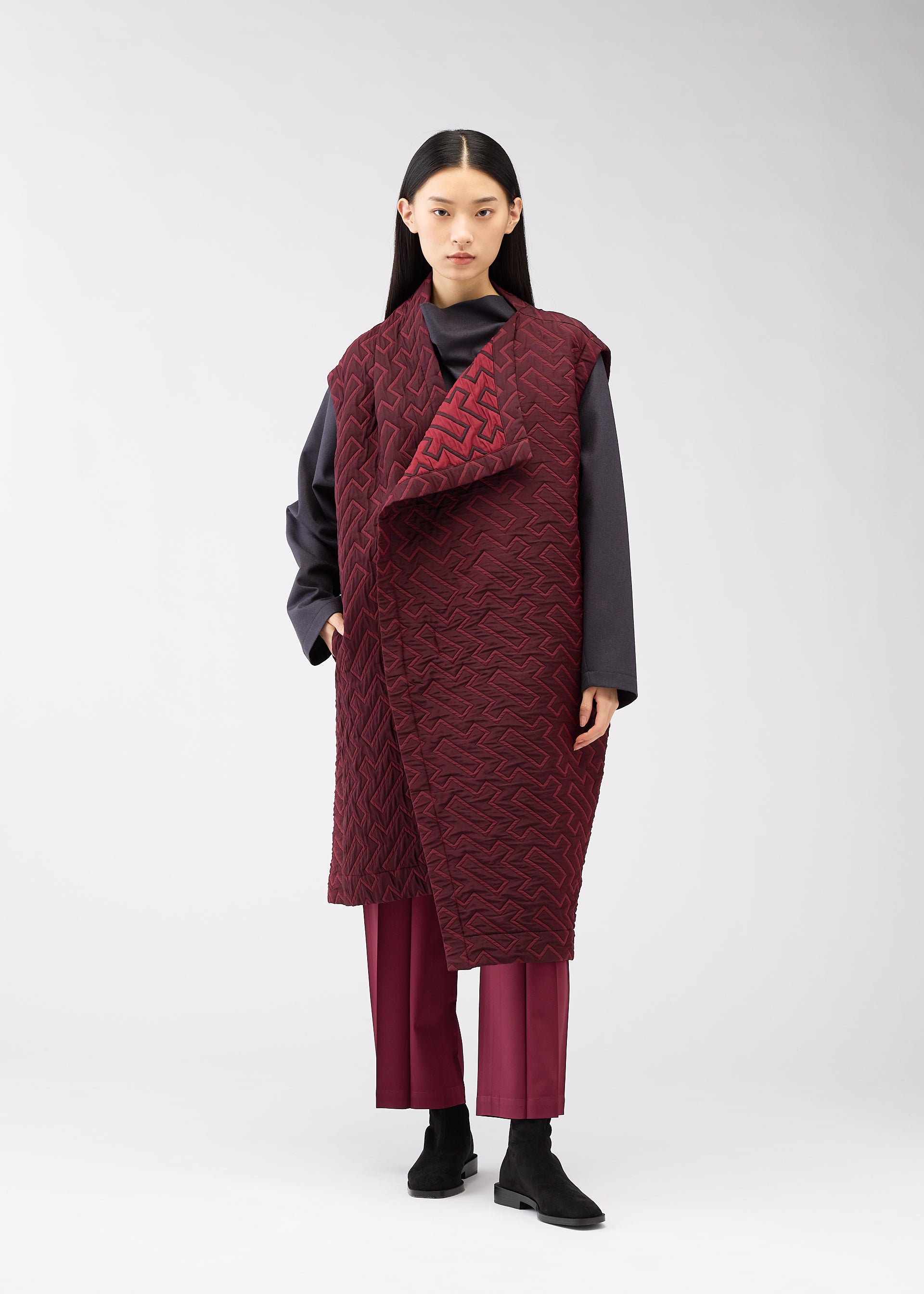 Zoomed view A person with long black hair wears the ISSEY MIYAKE ONLINE STORE's ONE CLOTH PUFF—a textured sleeveless maroon jacquard vest—over a dark gray top and maroon pants with black ankle boots, standing against a plain light background.