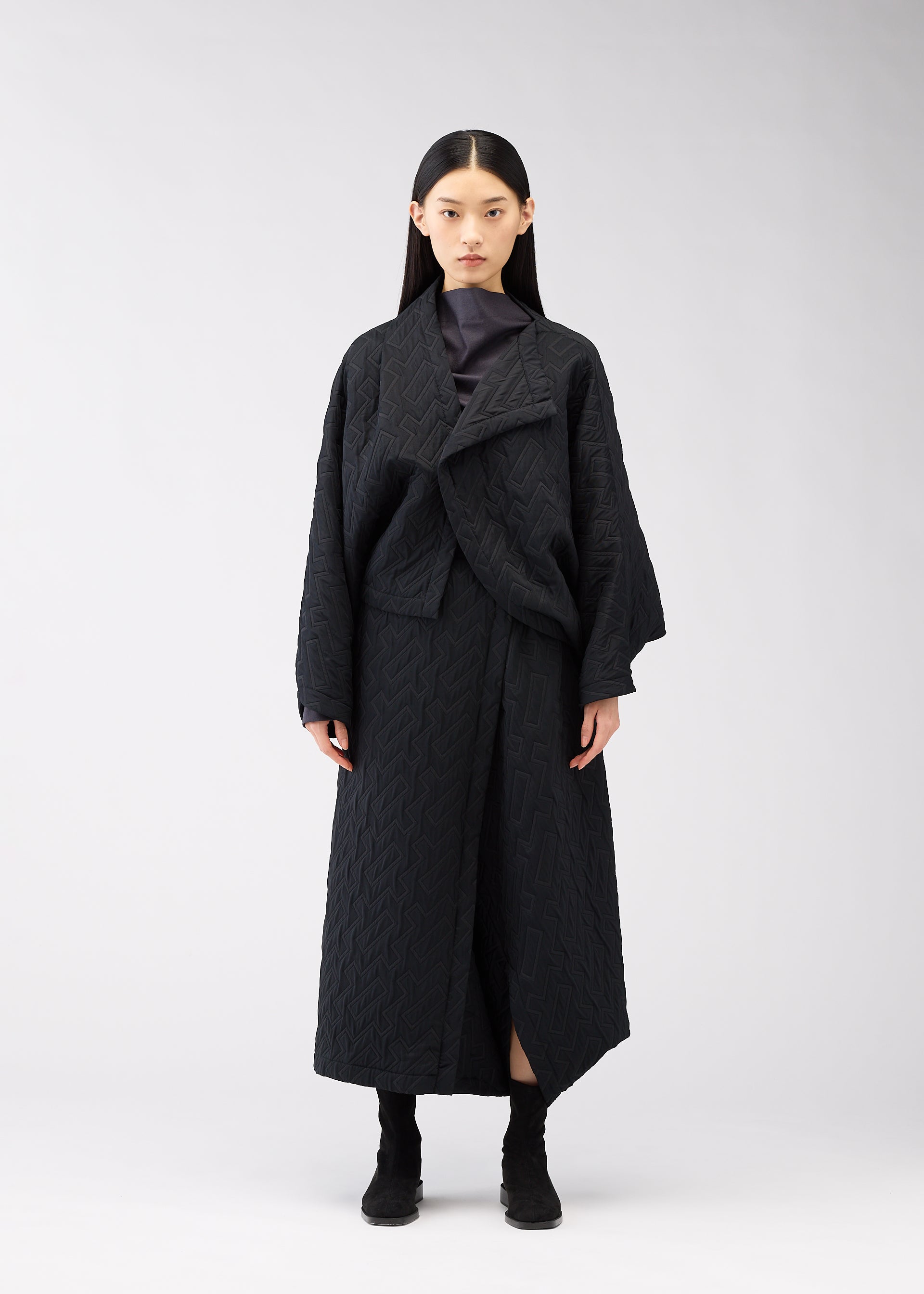 Zoomed view A woman with long straight black hair stands facing forward, wearing the ONE CLOTH PUFF jacket by 132 5. ISSEY MIYAKE over a dark top and black boots, against a plain white background.