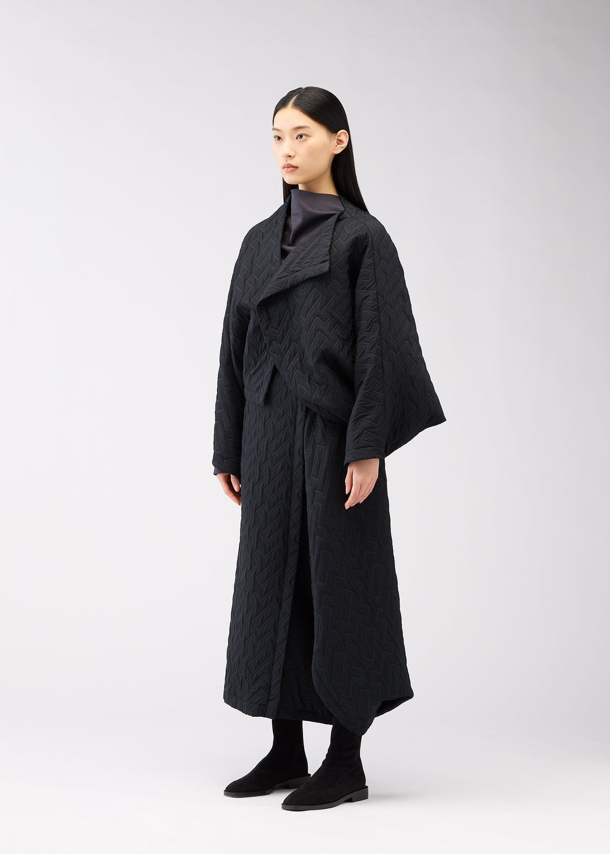 A woman with long, straight dark hair stands against a plain light background, wearing the ONE CLOTH PUFF jacket by 132 5. ISSEY MIYAKE, paired with black pants and black ankle boots, gazing slightly to the side. view 6 of 9