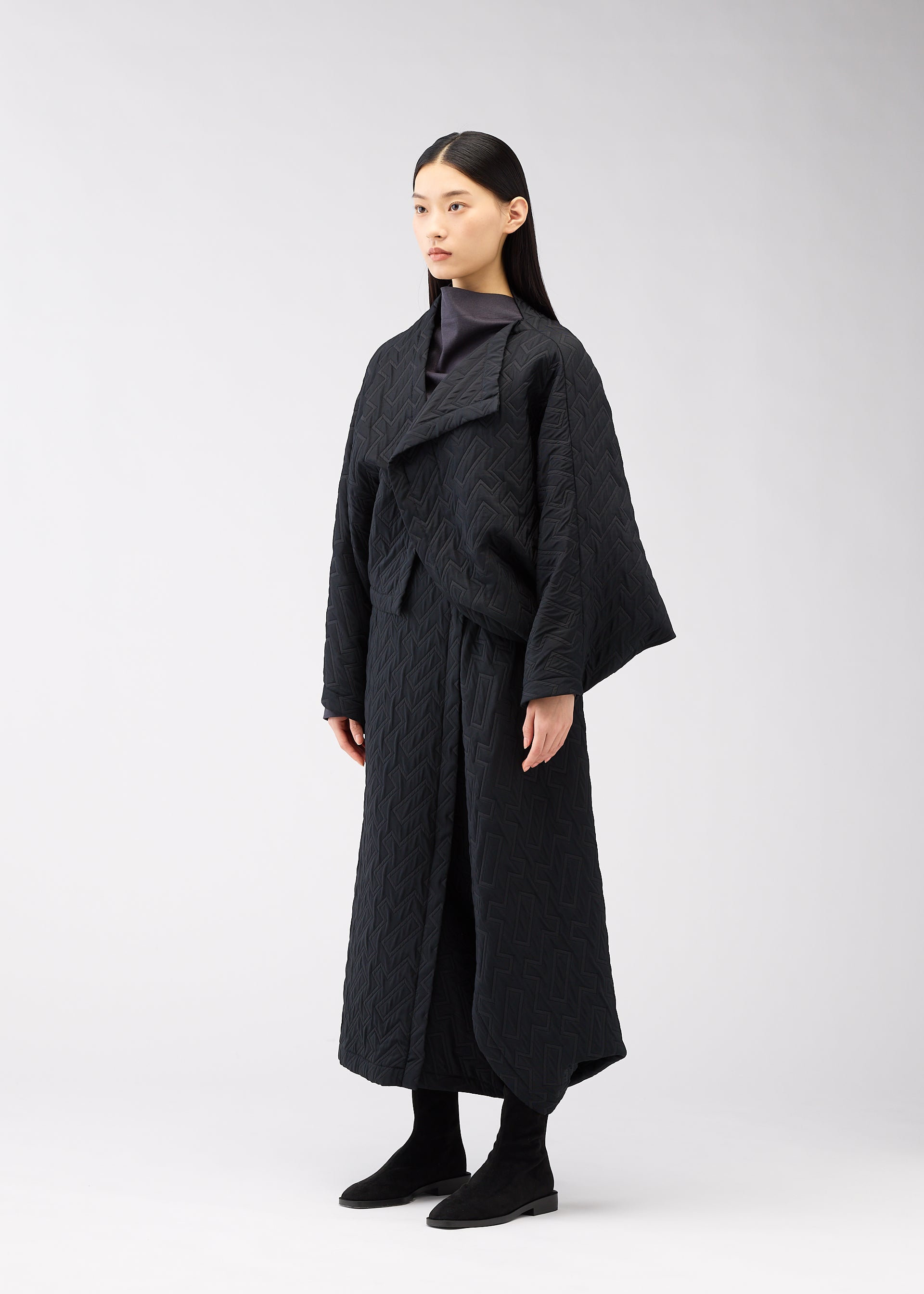 Zoomed view A woman with long, straight dark hair stands against a plain light background, wearing the ONE CLOTH PUFF jacket by 132 5. ISSEY MIYAKE, paired with black pants and black ankle boots, gazing slightly to the side.