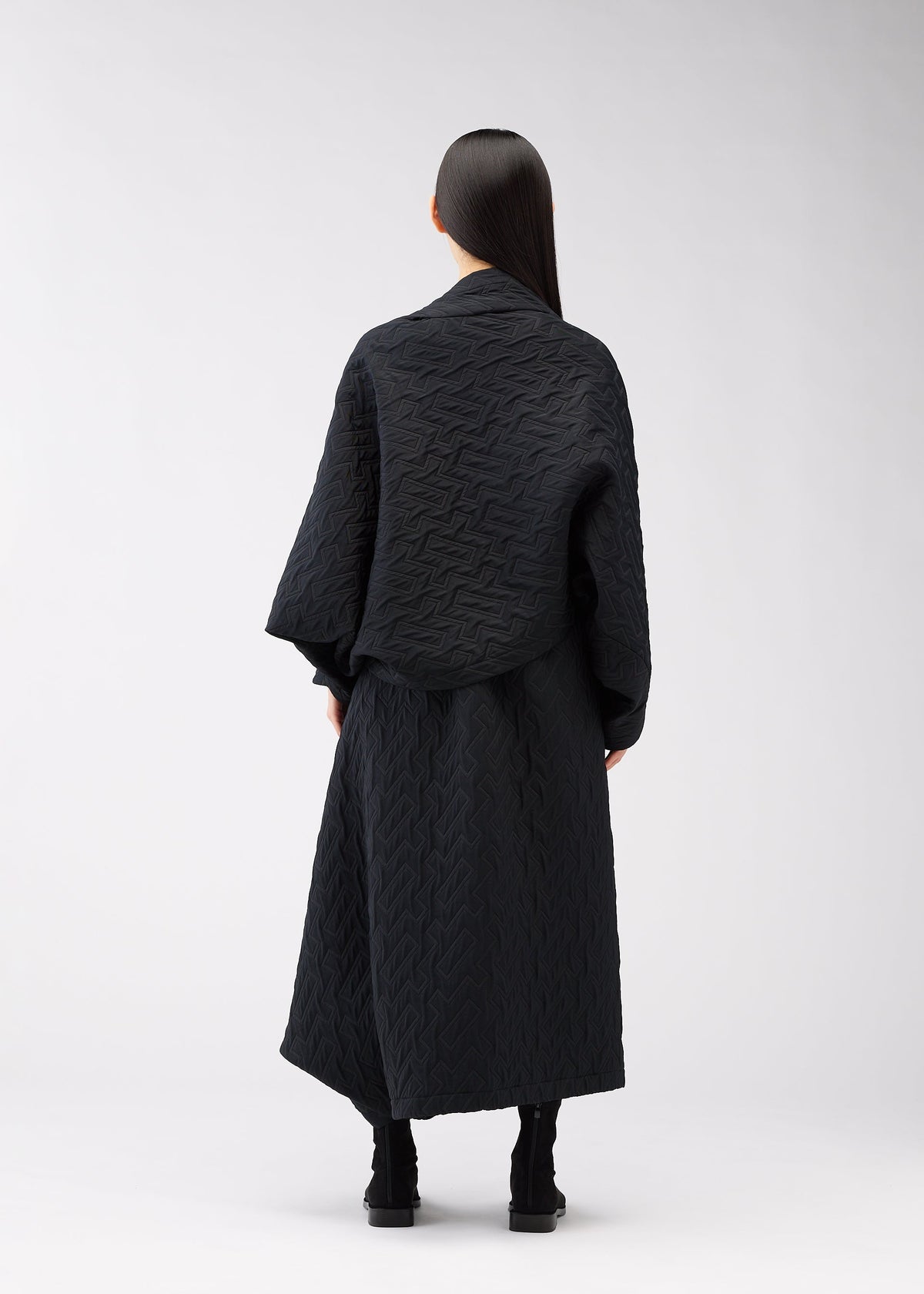 A person with long, straight dark hair stands facing away, wearing the ONE CLOTH PUFF jacquard weave jacket and a long skirt by 132 5. ISSEY MIYAKE, paired with black boots against a plain light background. view 7 of 9