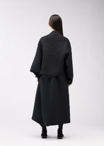 A person with long, straight dark hair stands facing away, wearing the ONE CLOTH PUFF jacquard weave jacket and a long skirt by 132 5. ISSEY MIYAKE, paired with black boots against a plain light background.