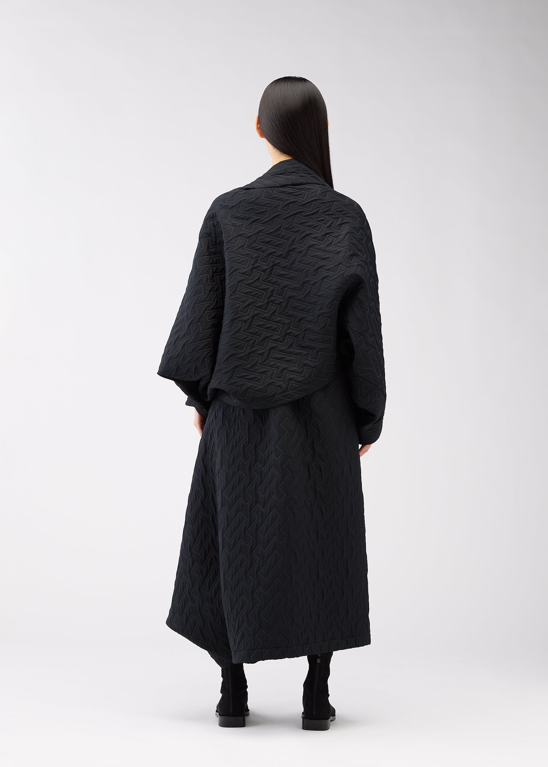 Zoomed view A person with long, straight dark hair stands facing away, wearing the ONE CLOTH PUFF jacquard weave jacket and a long skirt by 132 5. ISSEY MIYAKE, paired with black boots against a plain light background.