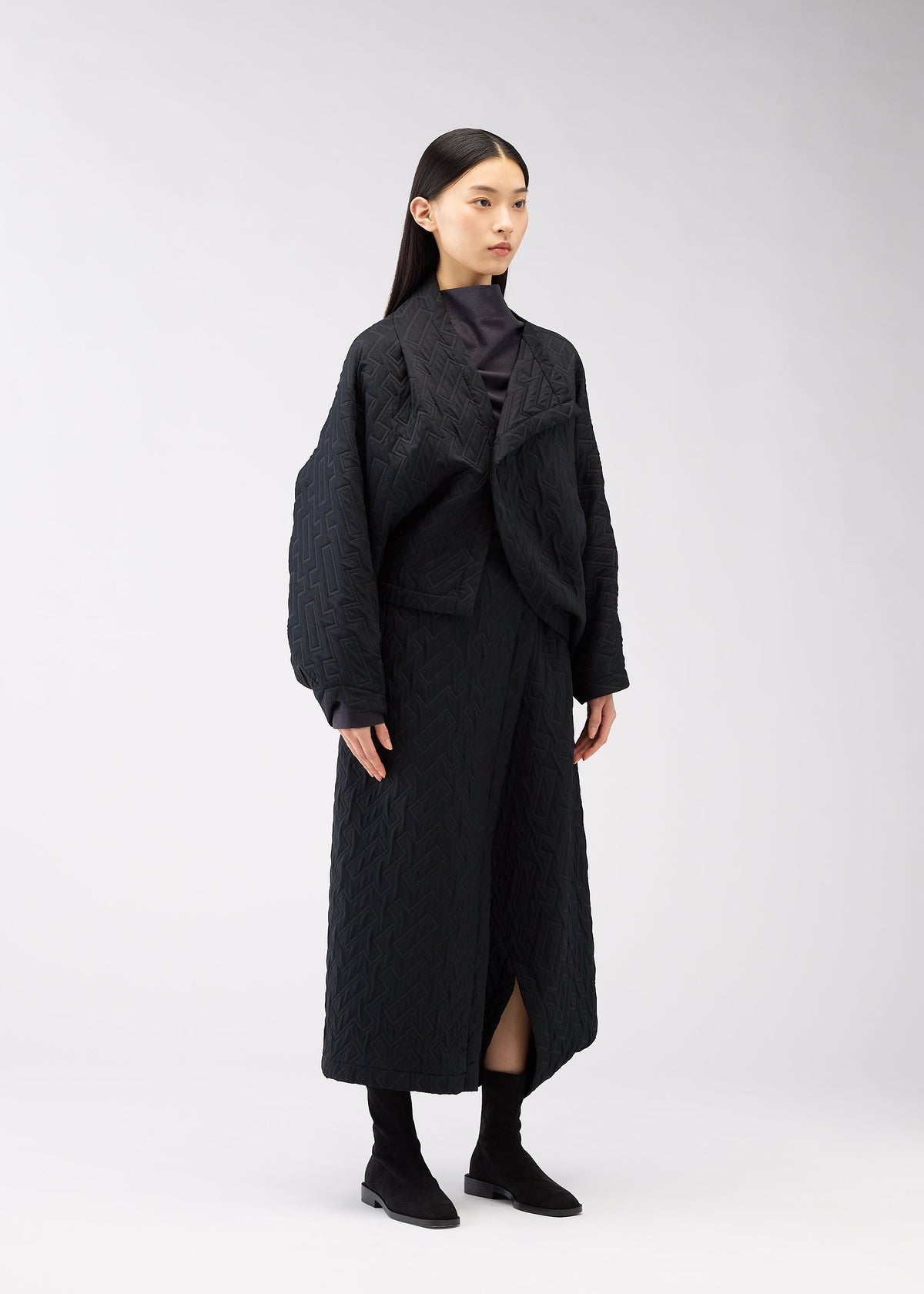 A woman with long black hair wears the ONE CLOTH PUFF jacquard weave jacket and matching long skirt from 132 5. ISSEY MIYAKE with black boots, standing against a plain light gray background. view 8 of 9