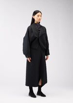 A woman with long black hair wears the ONE CLOTH PUFF jacquard weave jacket and matching long skirt from 132 5. ISSEY MIYAKE with black boots, standing against a plain light gray background.