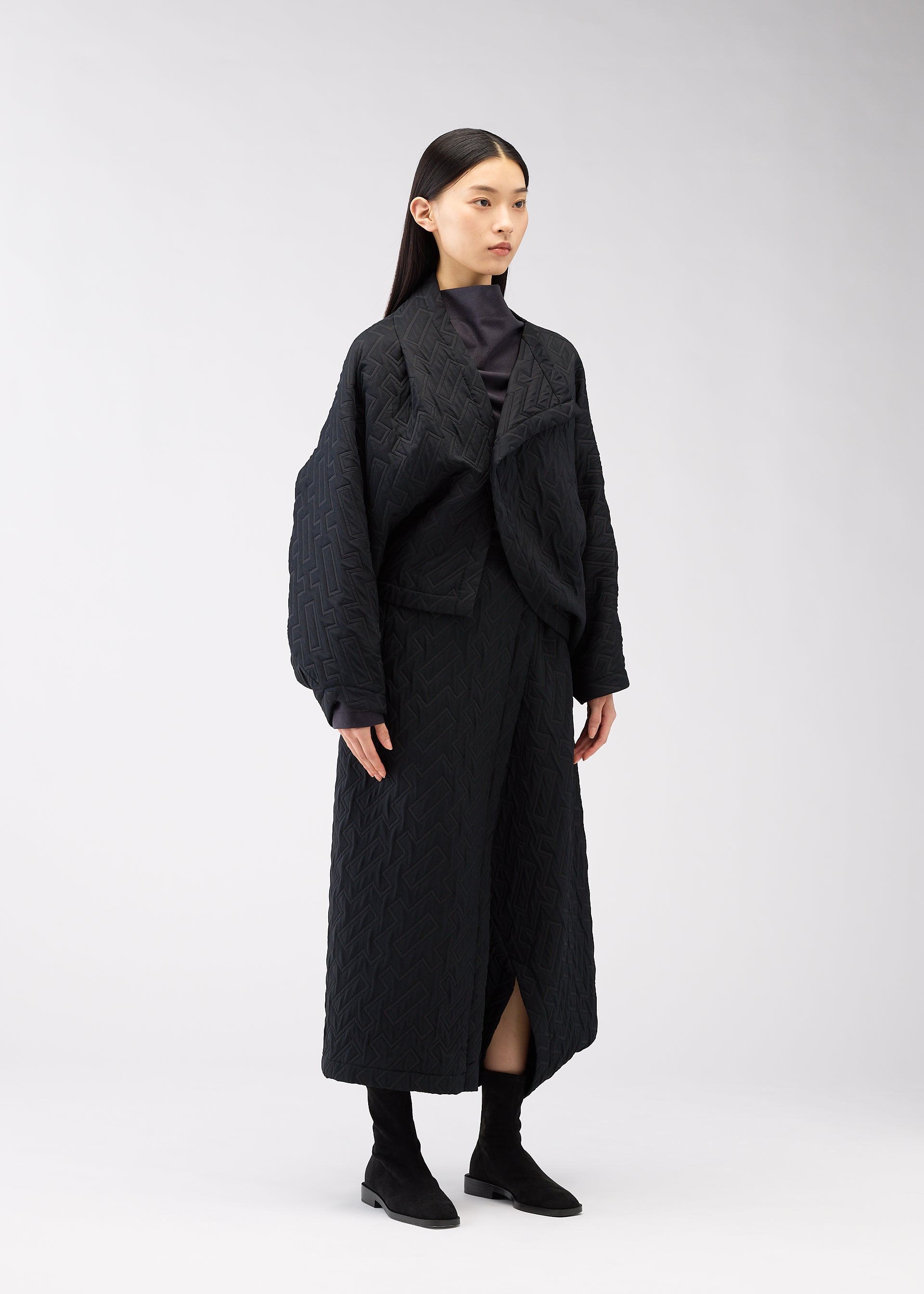 Zoomed view A woman with long black hair wears the ONE CLOTH PUFF jacquard weave jacket and matching long skirt from 132 5. ISSEY MIYAKE with black boots, standing against a plain light gray background.
