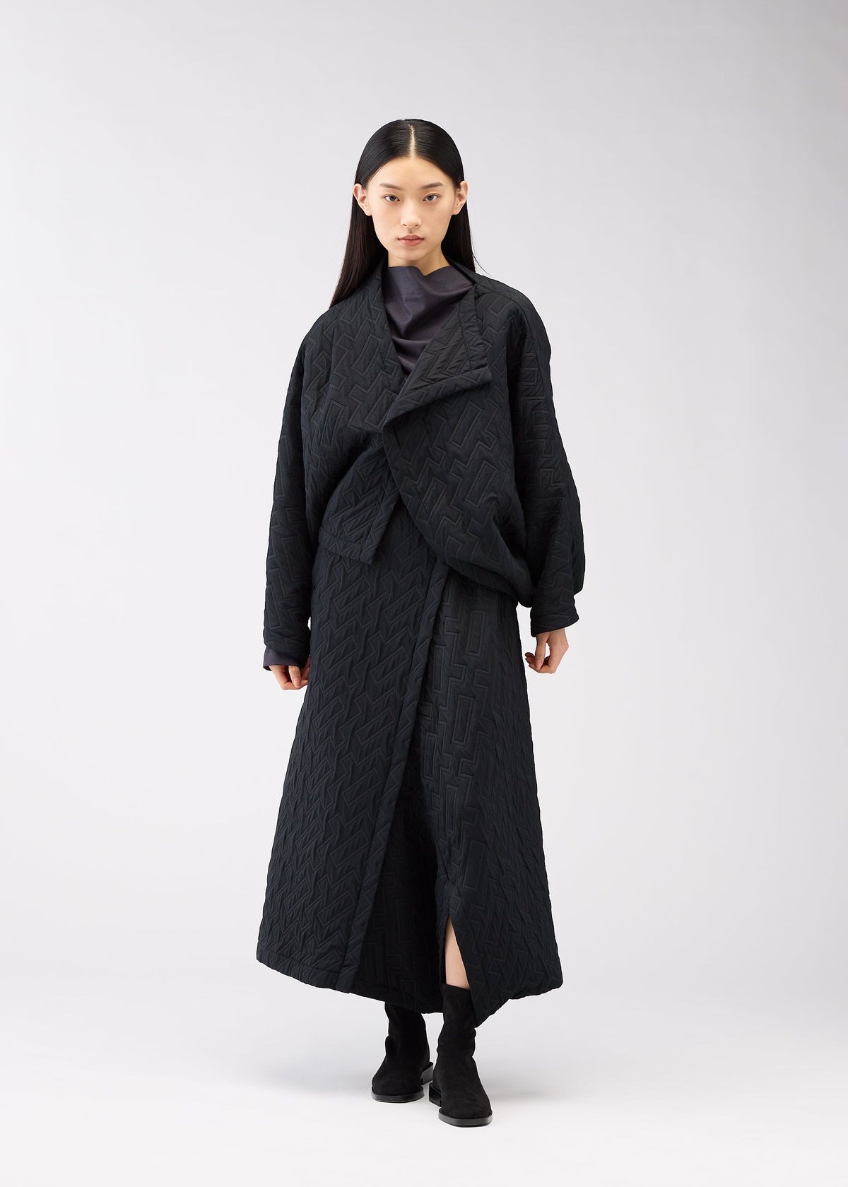 A person with loose straight dark hair stands against a light background, wearing the ONE CLOTH PUFF jacket by 132 5. ISSEY MIYAKE, paired with a textured black skirt and matching black boots, sporting a neutral expression. view 9 of 9