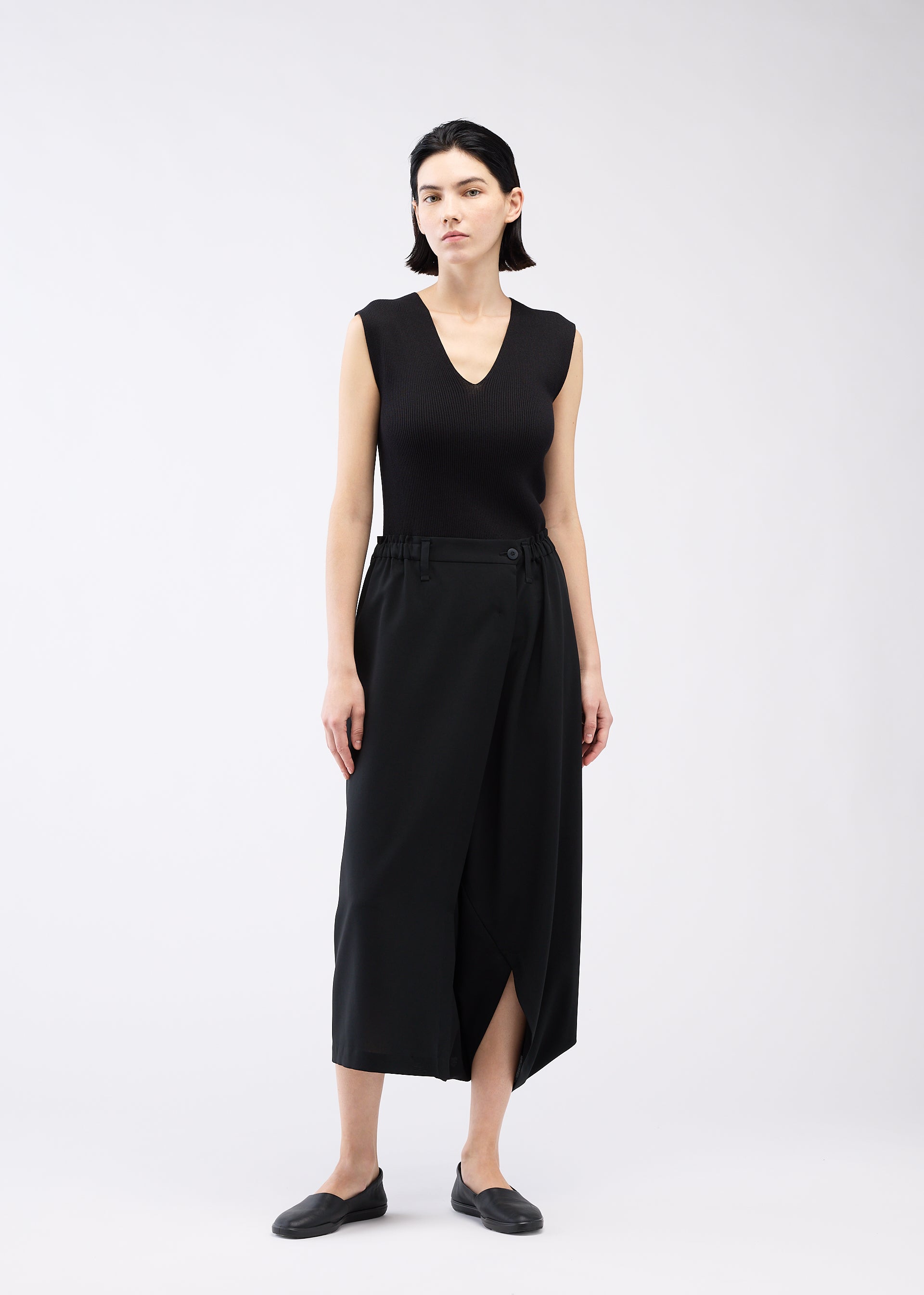 Zoomed view A woman with straight dark hair wears 132 5. ISSEY MIYAKE SEAMLESS BOTTOMS styled as skirt-like pants, with a sleeveless black V-neck top and black flats, standing against a plain white background.
