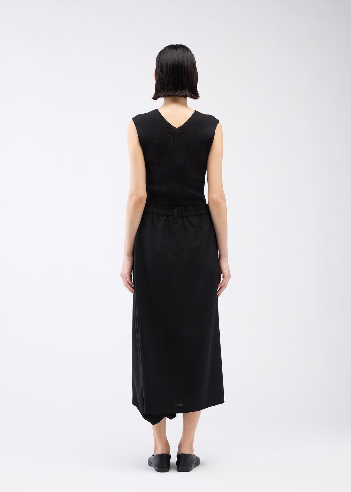 A person with short dark hair, seen from behind, wears a sleeveless black V-back top and SEAMLESS BOTTOMS by 132 5. ISSEY MIYAKE that resemble a long skirt. They stand against a plain white background in black slip-on shoes. view 10 of 11