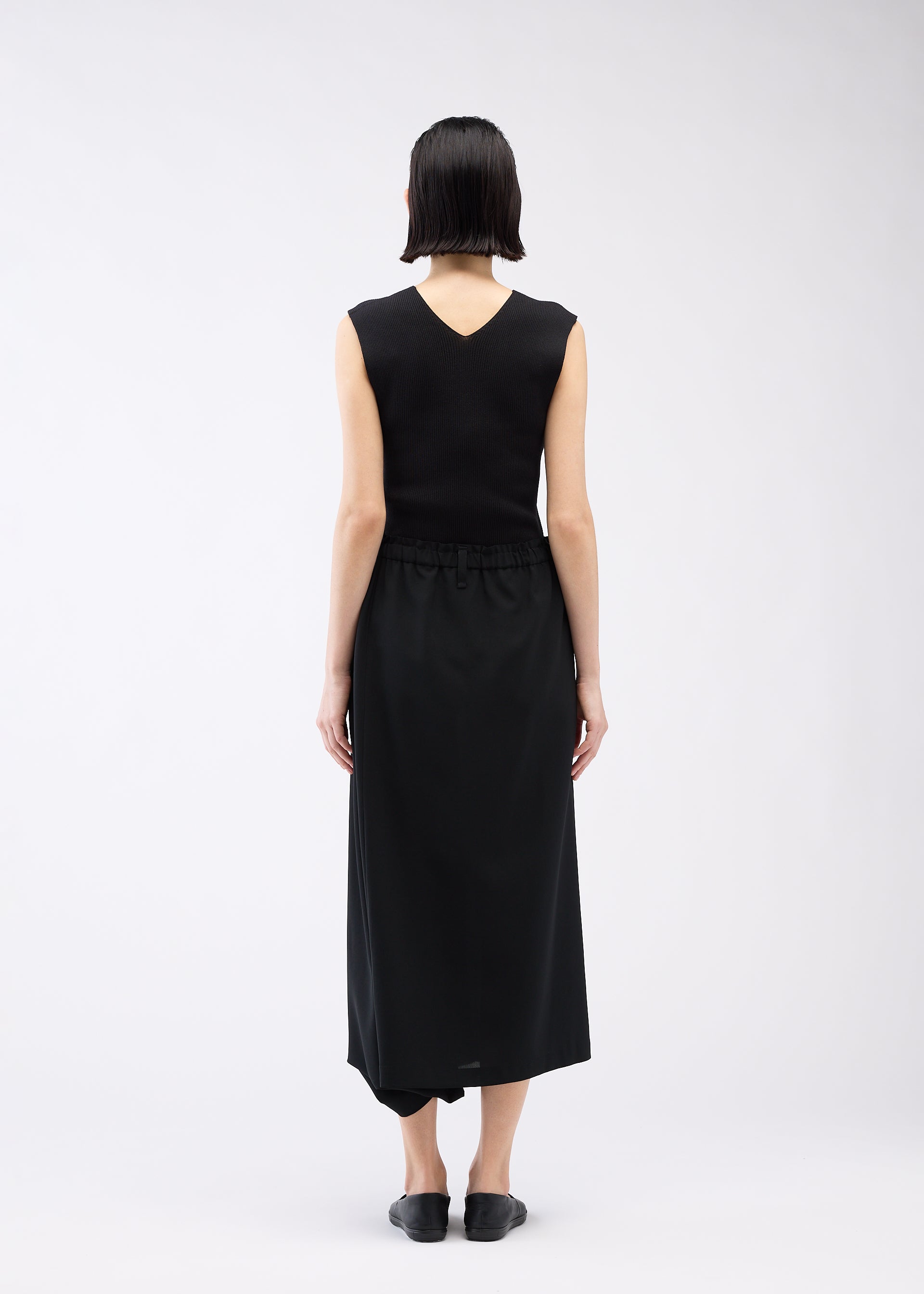 Zoomed view A person with short dark hair, seen from behind, wears a sleeveless black V-back top and SEAMLESS BOTTOMS by 132 5. ISSEY MIYAKE that resemble a long skirt. They stand against a plain white background in black slip-on shoes.