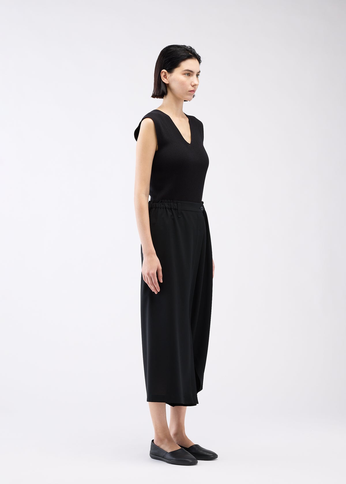 A woman with short dark hair, wearing 132 5. ISSEY MIYAKE’s SEAMLESS BOTTOMS and a sleeveless black V-neck top, stands sideways in black slip-on shoes against a plain light background. view 11 of 11