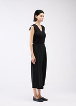 A woman with short dark hair, wearing 132 5. ISSEY MIYAKE’s SEAMLESS BOTTOMS and a sleeveless black V-neck top, stands sideways in black slip-on shoes against a plain light background.