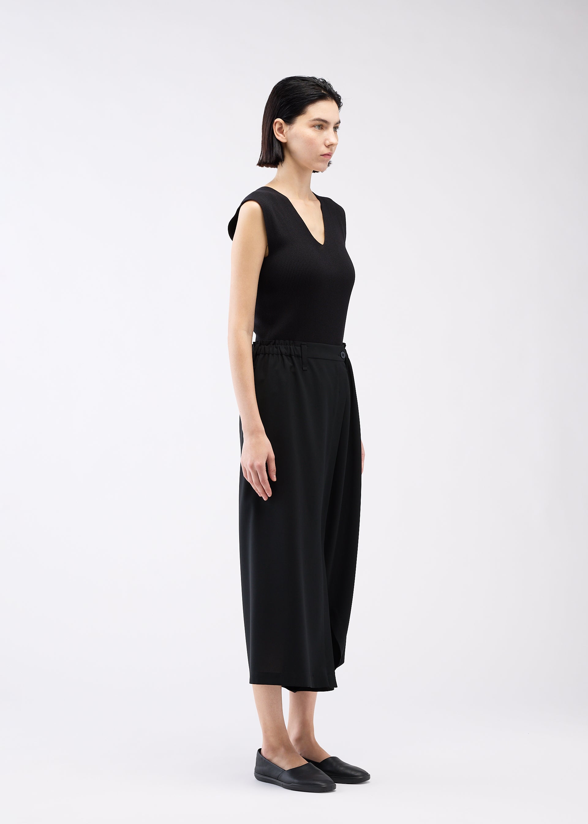 Zoomed view A woman with short dark hair, wearing 132 5. ISSEY MIYAKE’s SEAMLESS BOTTOMS and a sleeveless black V-neck top, stands sideways in black slip-on shoes against a plain light background.