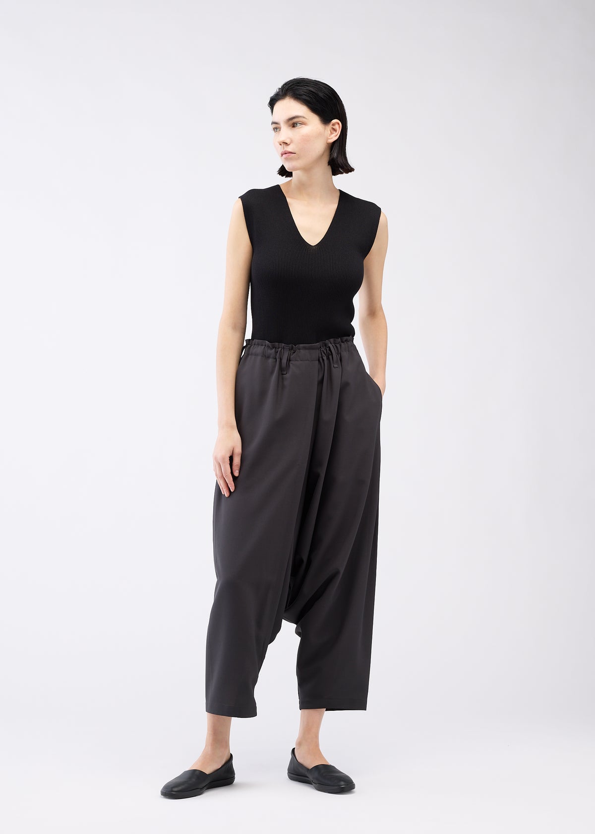 A woman with short dark hair models the SEAMLESS BOTTOMS by 132 5. ISSEY MIYAKE—loose, high-waisted sarouel pants with side pockets and a relaxed silhouette—paired with a sleeveless black top against a plain light background. view 9 of 12