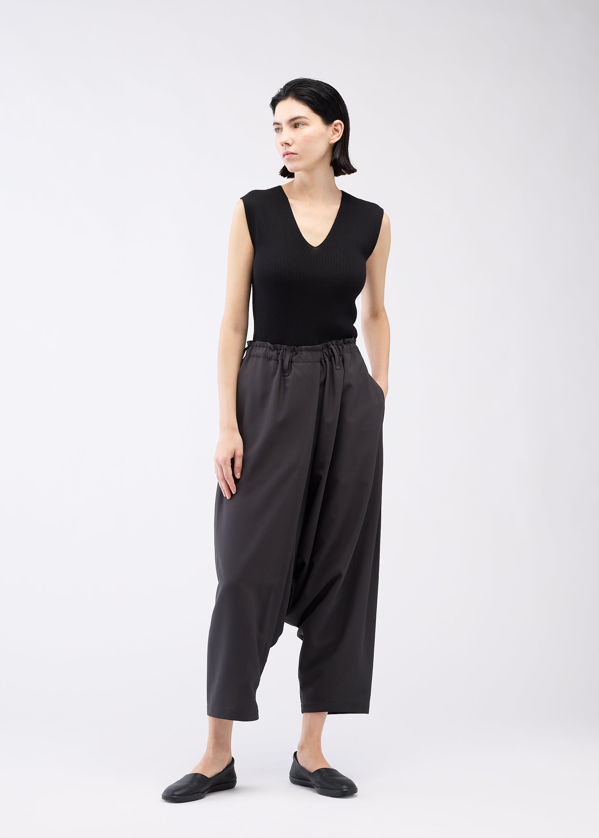 Zoomed view A woman with short dark hair models the SEAMLESS BOTTOMS by 132 5. ISSEY MIYAKE—loose, high-waisted sarouel pants with side pockets and a relaxed silhouette—paired with a sleeveless black top against a plain light background.