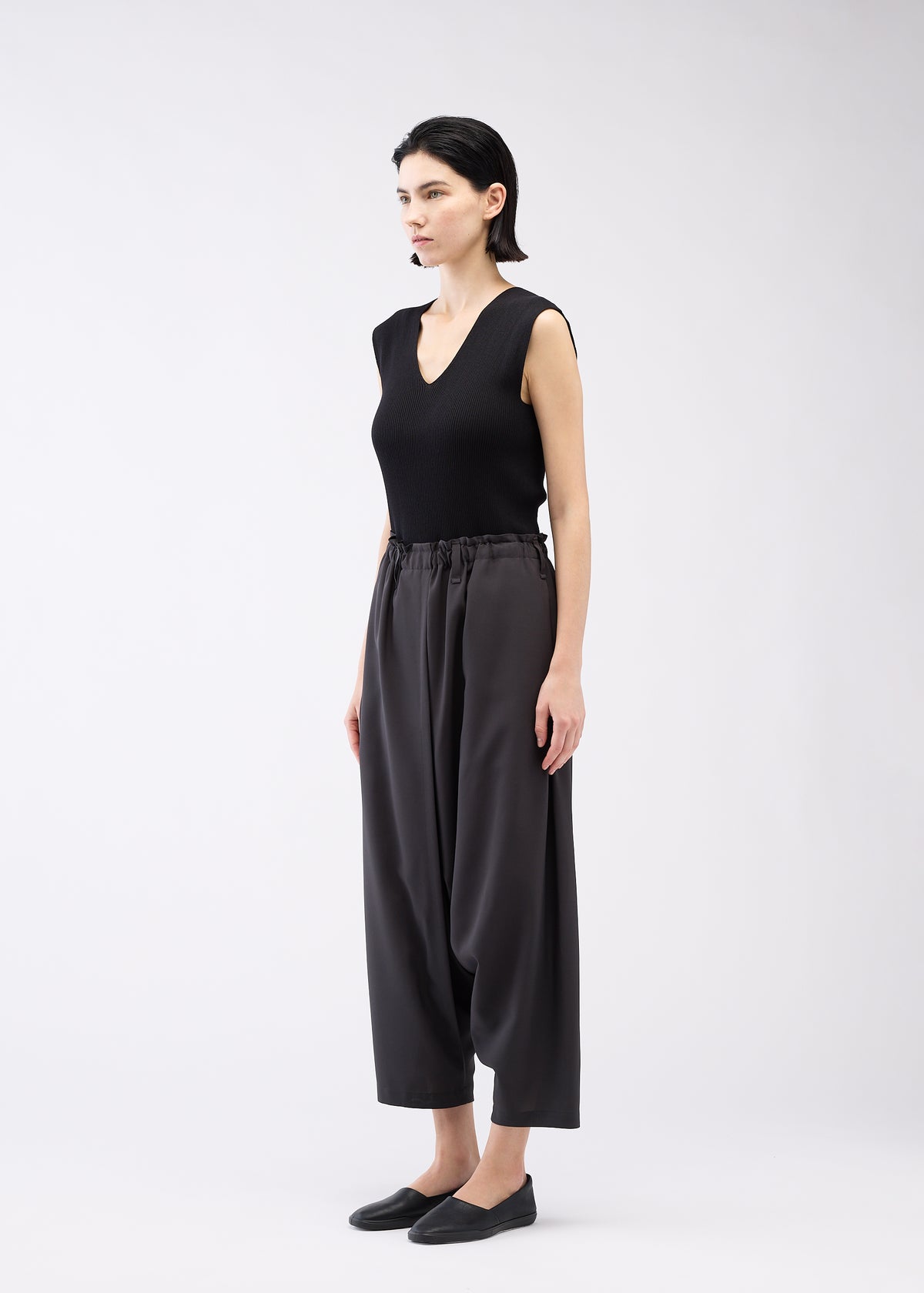 A woman with short dark hair and a neutral expression stands against a white background, wearing 132 5. ISSEY MIYAKE SEAMLESS BOTTOMS, a sleeveless black V-neck top, and black slip-on shoes for a relaxed silhouette. view 11 of 12