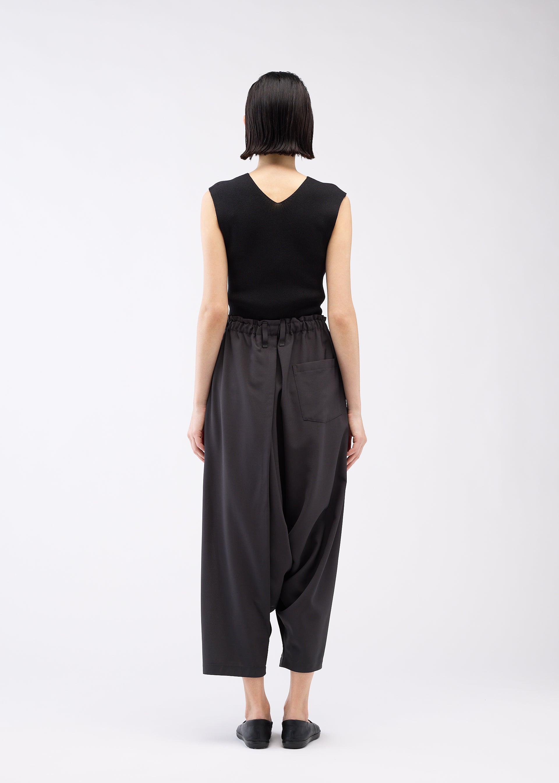 Zoomed view A person with short dark hair, wearing a sleeveless black top and 132 5. ISSEY MIYAKE SEAMLESS BOTTOMS with a relaxed silhouette, stands facing away from the camera against a plain white background.