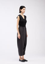 A woman with short dark hair stands sideways against a plain white background, wearing a sleeveless black top, black slip-on shoes, and SEAMLESS BOTTOMS by 132 5. ISSEY MIYAKE made from recycled polyester with a relaxed silhouette.