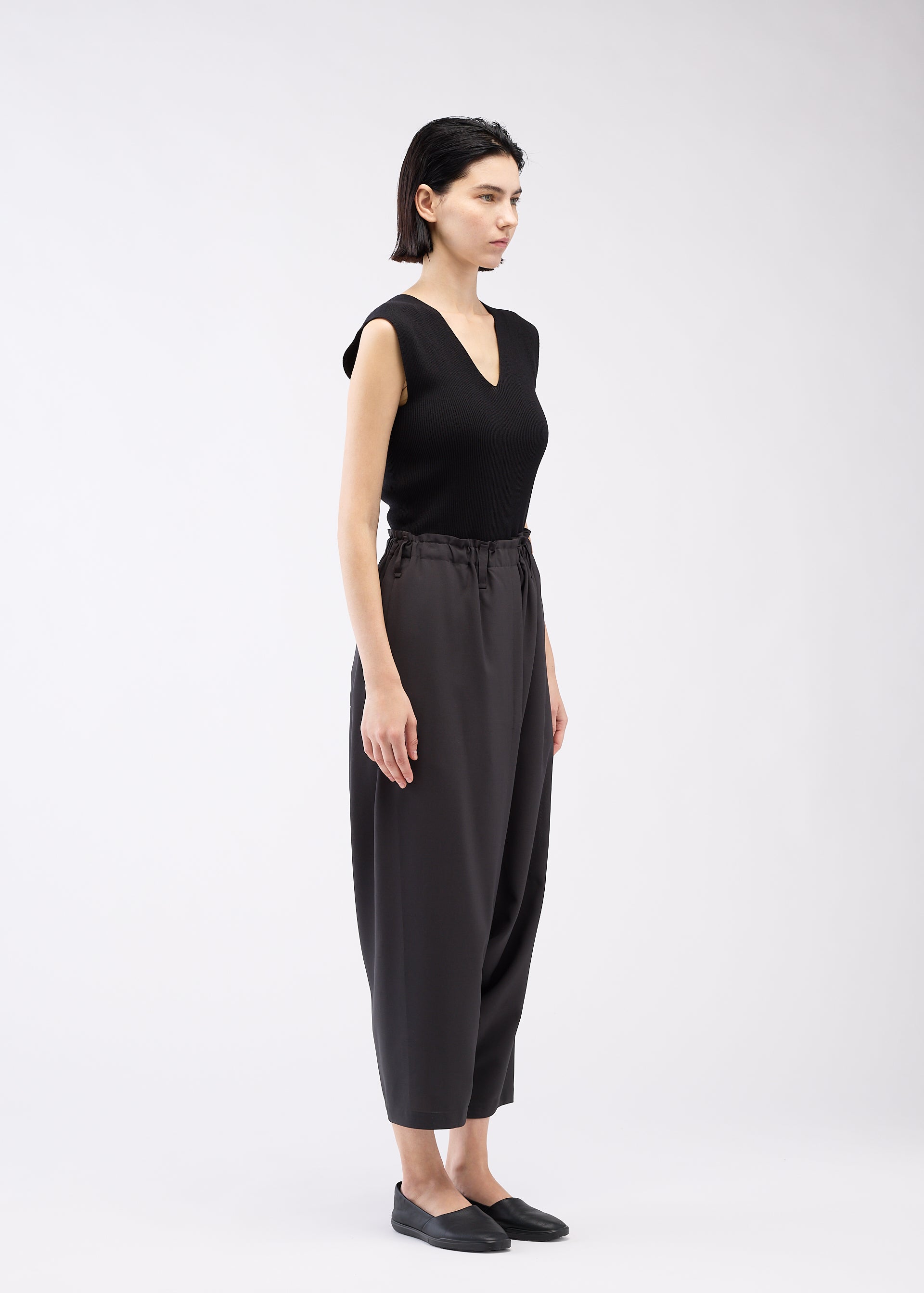 Zoomed view A woman with short dark hair stands sideways against a plain white background, wearing a sleeveless black top, black slip-on shoes, and SEAMLESS BOTTOMS by 132 5. ISSEY MIYAKE made from recycled polyester with a relaxed silhouette.