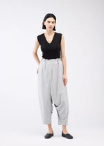 A person with short dark hair wears a sleeveless black V-neck top, black flat shoes, and loose high-waisted JERSEY BOTTOMS BASIC by 132 5. ISSEY MIYAKE, standing against a plain white background.