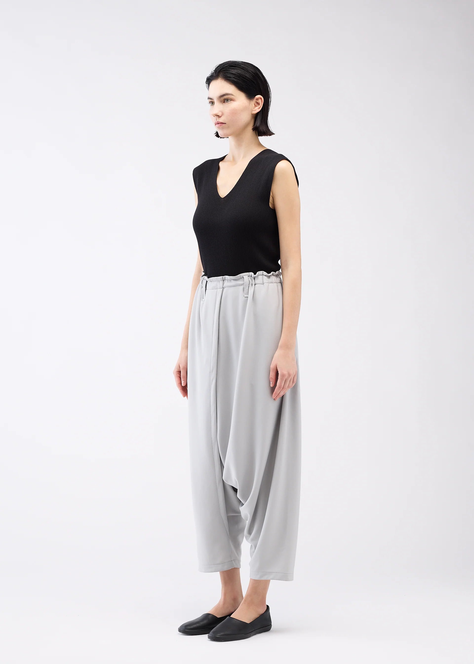 Zoomed view A person stands against a plain white background, wearing 132 5. ISSEY MIYAKE JERSEY BOTTOMS BASIC in loose light gray for a relaxed silhouette, paired with a sleeveless black top and black slip-on shoes, facing slightly left with a neutral expression.