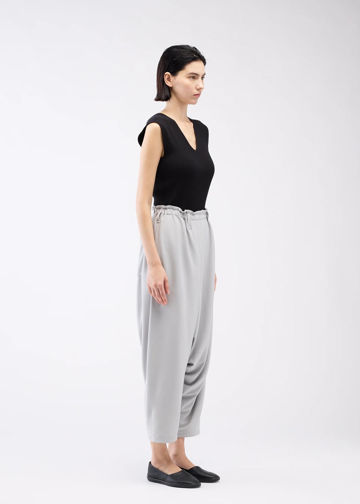 A woman stands in profile against a plain light background, wearing a sleeveless black top, black slip-on shoes, and light gray JERSEY BOTTOMS BASIC by 132 5. ISSEY MIYAKE—high-waisted Sarouel pants with an elastic waistband for a relaxed look. view 10 of 11