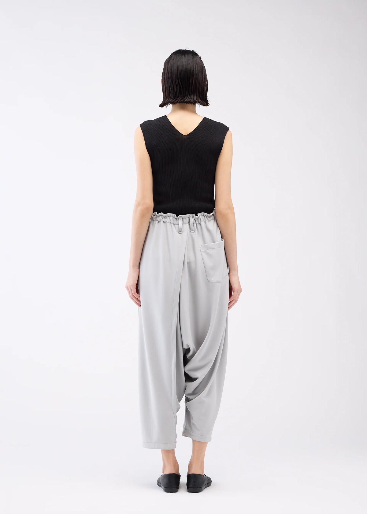 A person with short dark hair, seen from behind, wears a sleeveless black top and JERSEY BOTTOMS BASIC by 132 5. ISSEY MIYAKE—light gray, relaxed silhouette pants with an elastic waist and pocket—plus black slip-ons against a white background. view 11 of 11