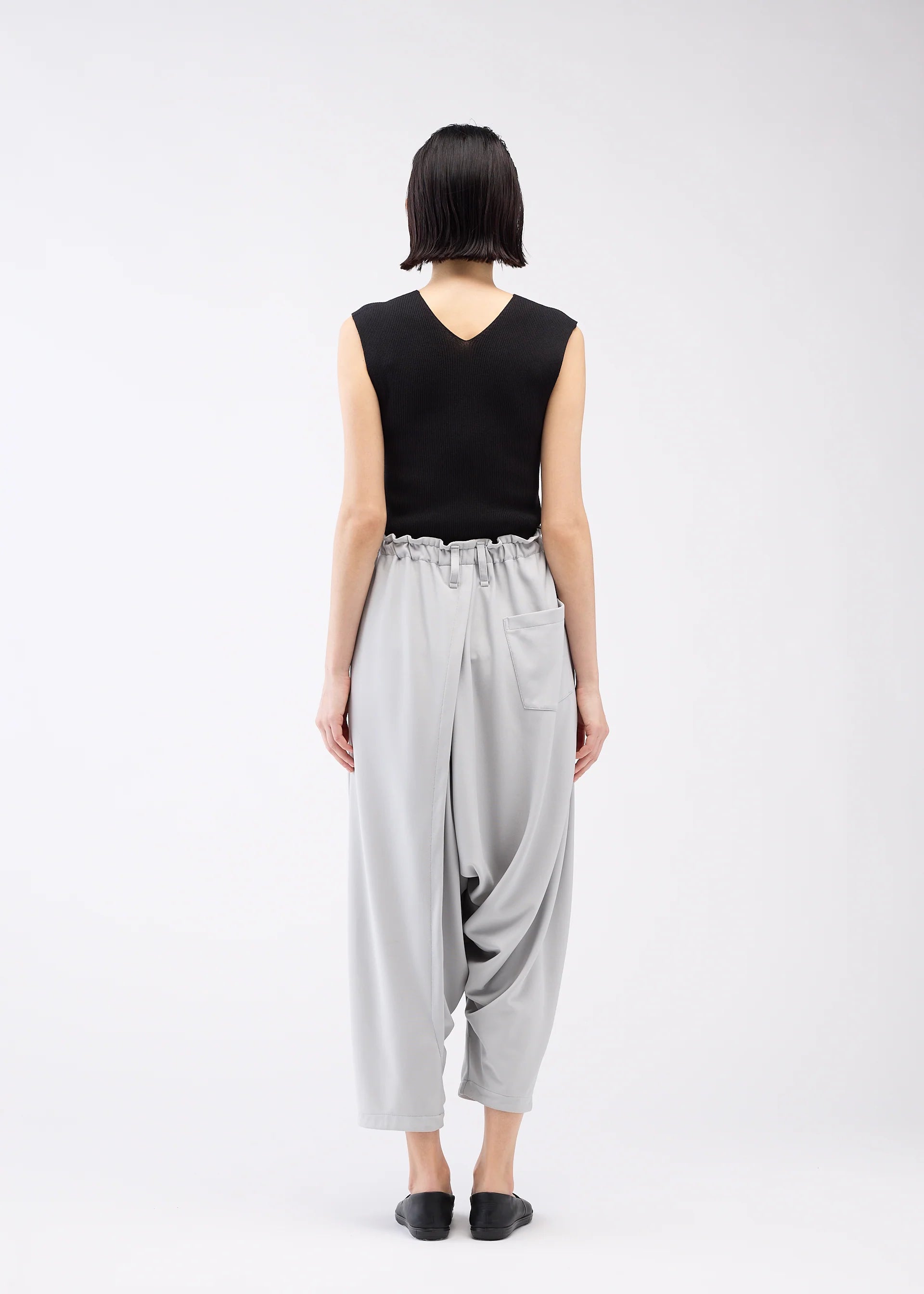Zoomed view A person with short dark hair, seen from behind, wears a sleeveless black top and JERSEY BOTTOMS BASIC by 132 5. ISSEY MIYAKE—light gray, relaxed silhouette pants with an elastic waist and pocket—plus black slip-ons against a white background.