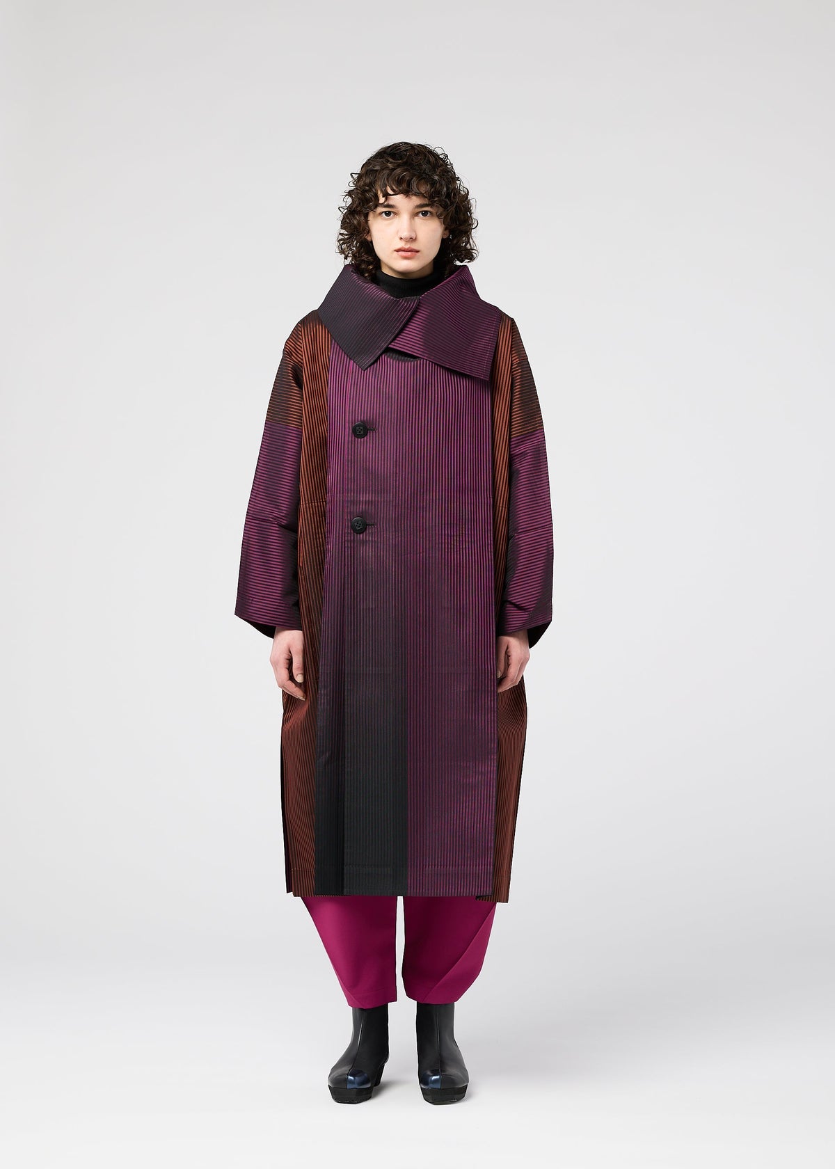 A person with curly hair wears the 132 5. ISSEY MIYAKE GRADATION COAT—an oversized jacquard coat in purple and brown with a large collar and big buttons—paired with magenta pants and black boots, standing against a plain white background. view 6 of 11
