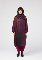 A person with curly hair wears the 132 5. ISSEY MIYAKE GRADATION COAT—an oversized jacquard coat in purple and brown with a large collar and big buttons—paired with magenta pants and black boots, standing against a plain white background.