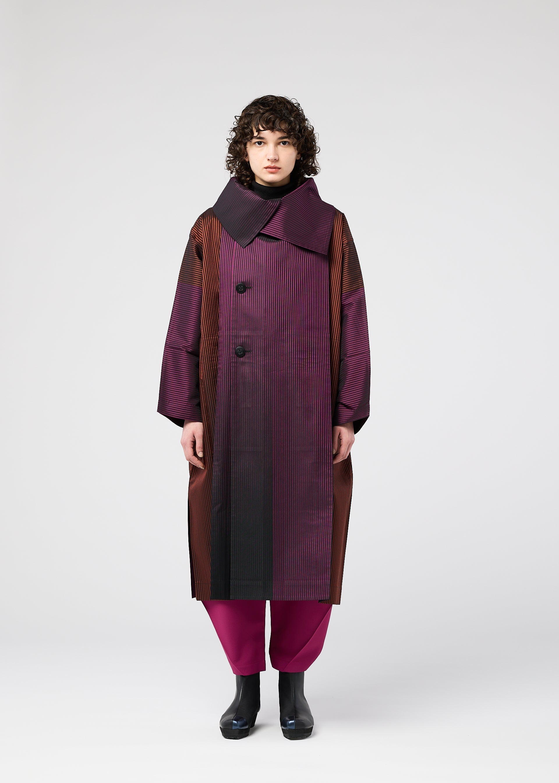Zoomed view A person with curly hair wears the 132 5. ISSEY MIYAKE GRADATION COAT—an oversized jacquard coat in purple and brown with a large collar and big buttons—paired with magenta pants and black boots, standing against a plain white background.