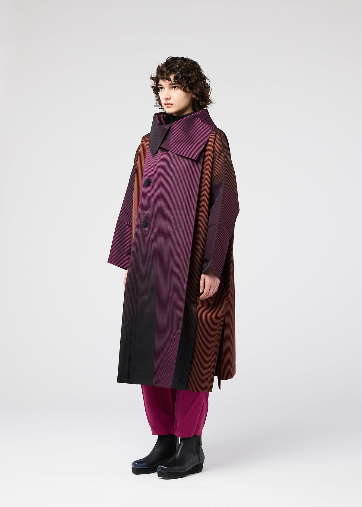 A person with curly hair models the GRADATION COAT by 132 5. ISSEY MIYAKE—an oversized jacquard coat in burgundy and brown with a large collar—paired with magenta pants and black boots, against a plain light background. view 7 of 11