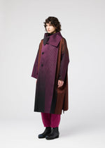 A person with curly hair models the GRADATION COAT by 132 5. ISSEY MIYAKE—an oversized jacquard coat in burgundy and brown with a large collar—paired with magenta pants and black boots, against a plain light background.