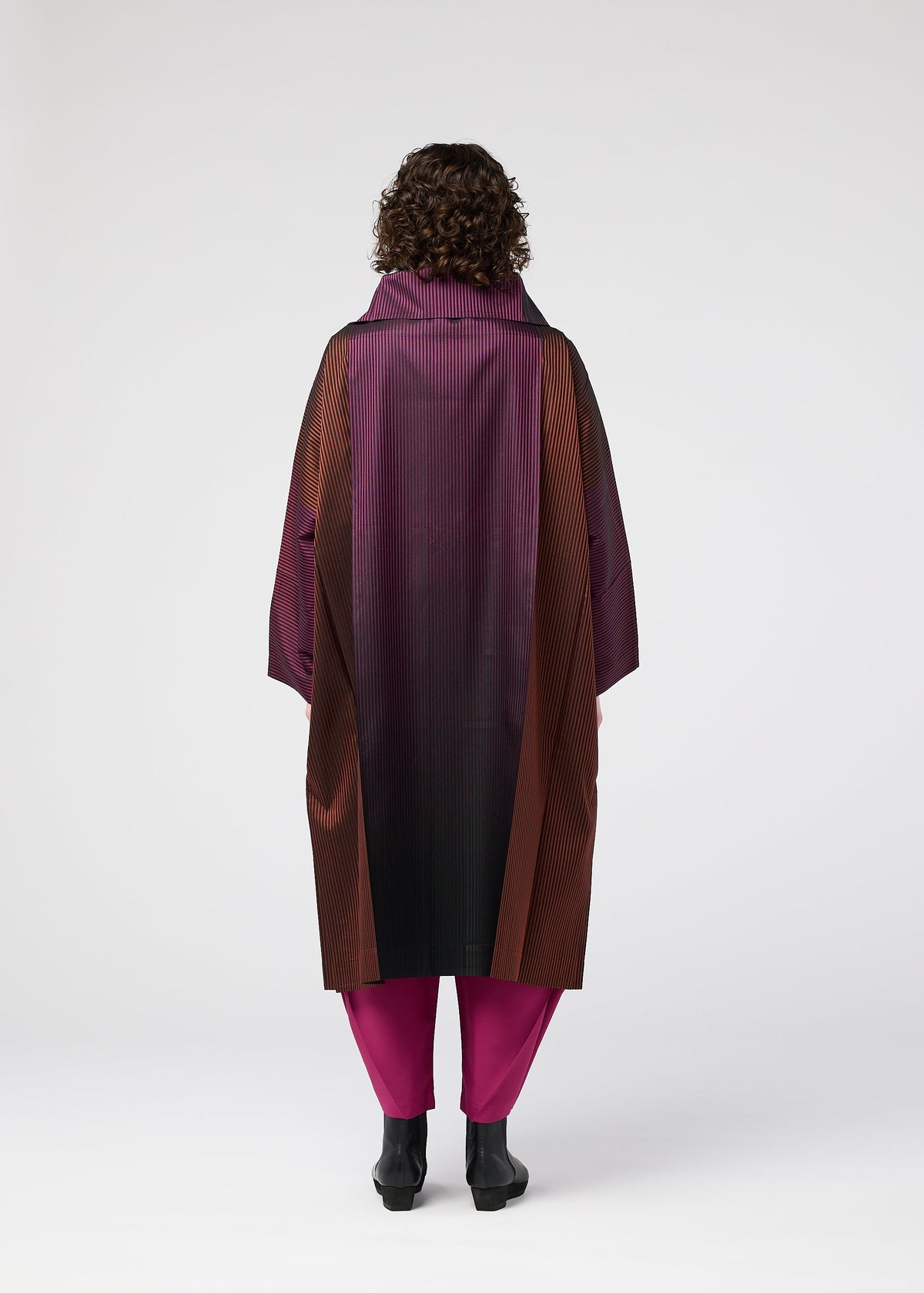 A person with curly hair stands facing away, wearing the GRADATION COAT by 132 5. ISSEY MIYAKE in purple, burgundy, and black with a large collar, paired with magenta pants and black shoes, against a plain white background. view 8 of 11