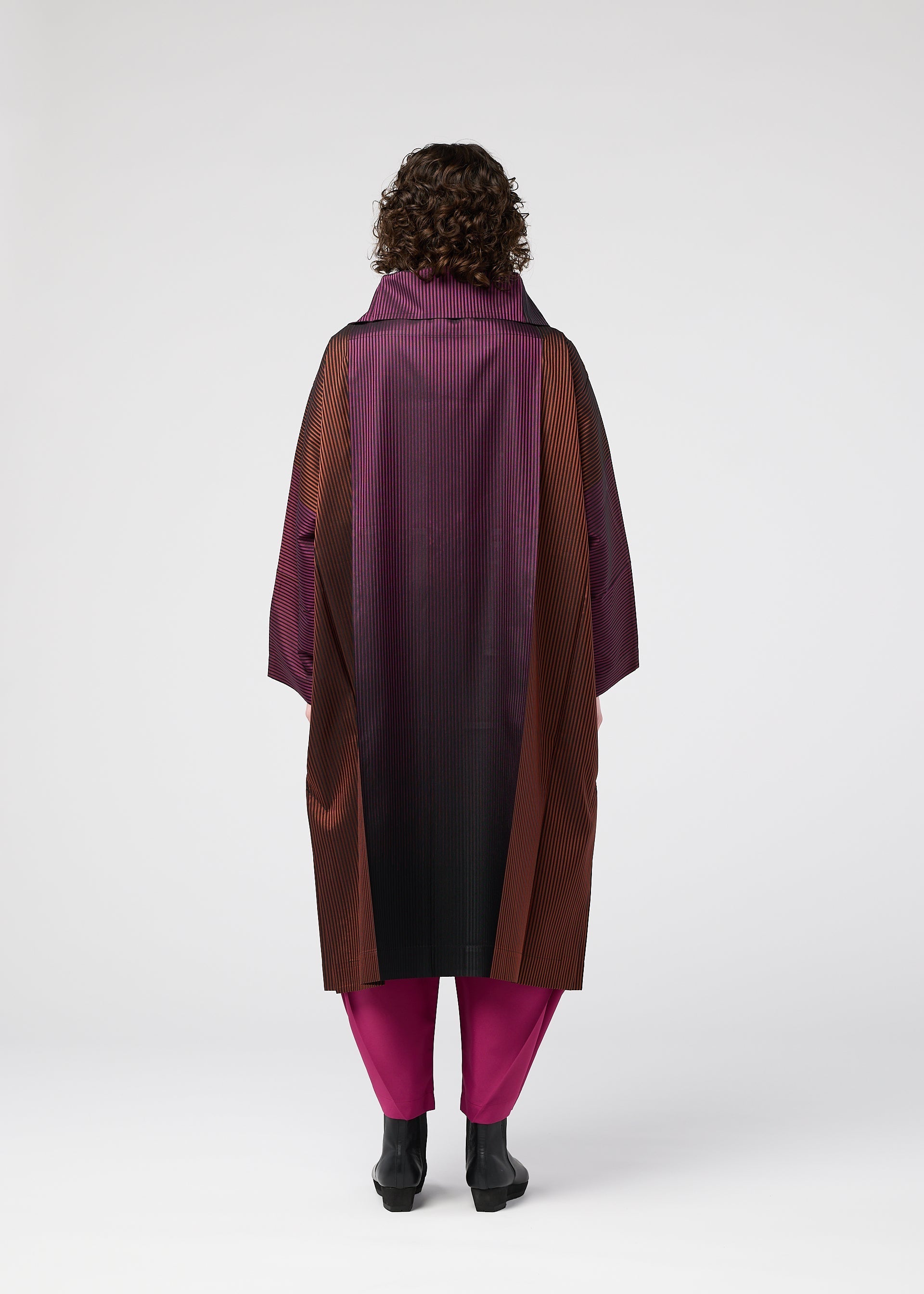 Zoomed view A person with curly hair stands facing away, wearing the GRADATION COAT by 132 5. ISSEY MIYAKE in purple, burgundy, and black with a large collar, paired with magenta pants and black shoes, against a plain white background.