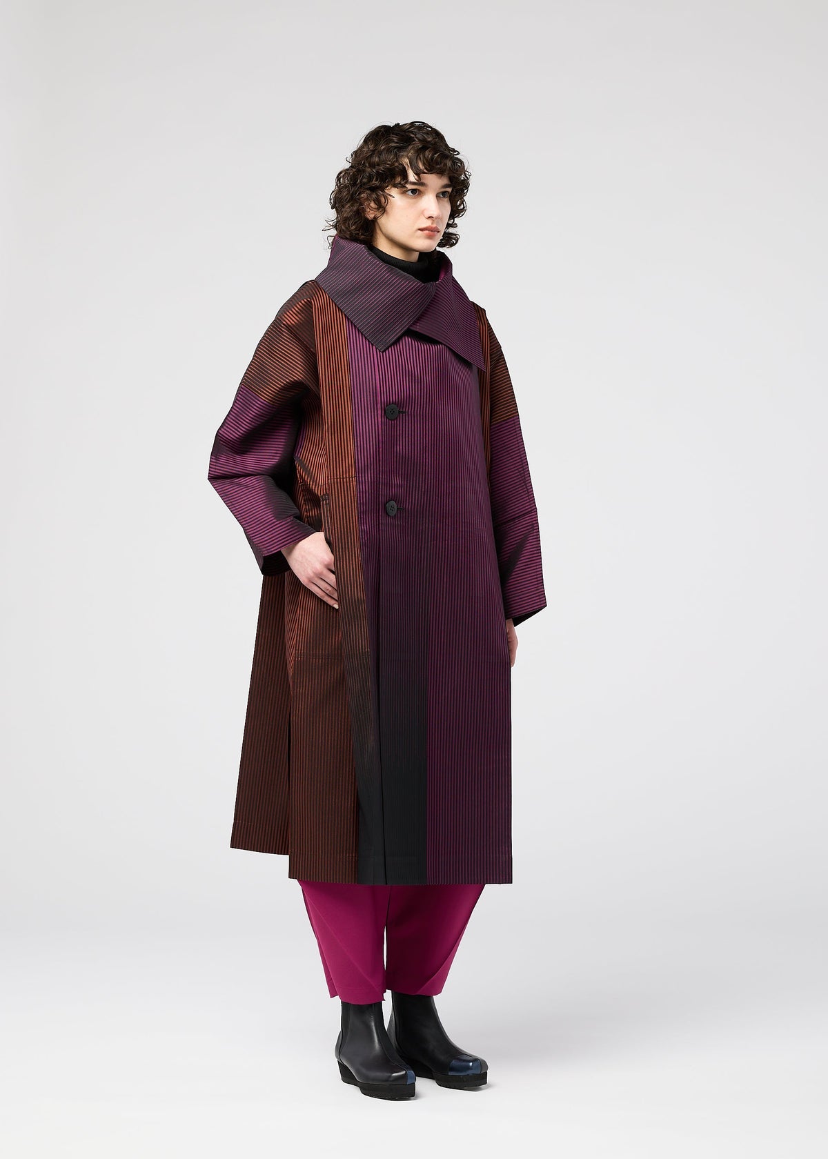 A person with curly hair models the GRADATION COAT by 132 5. ISSEY MIYAKE, a long, oversized jacquard coat with vertical stripes in burgundy, brown, and black. Styled with magenta pants and black boots against a plain white background. view 9 of 11