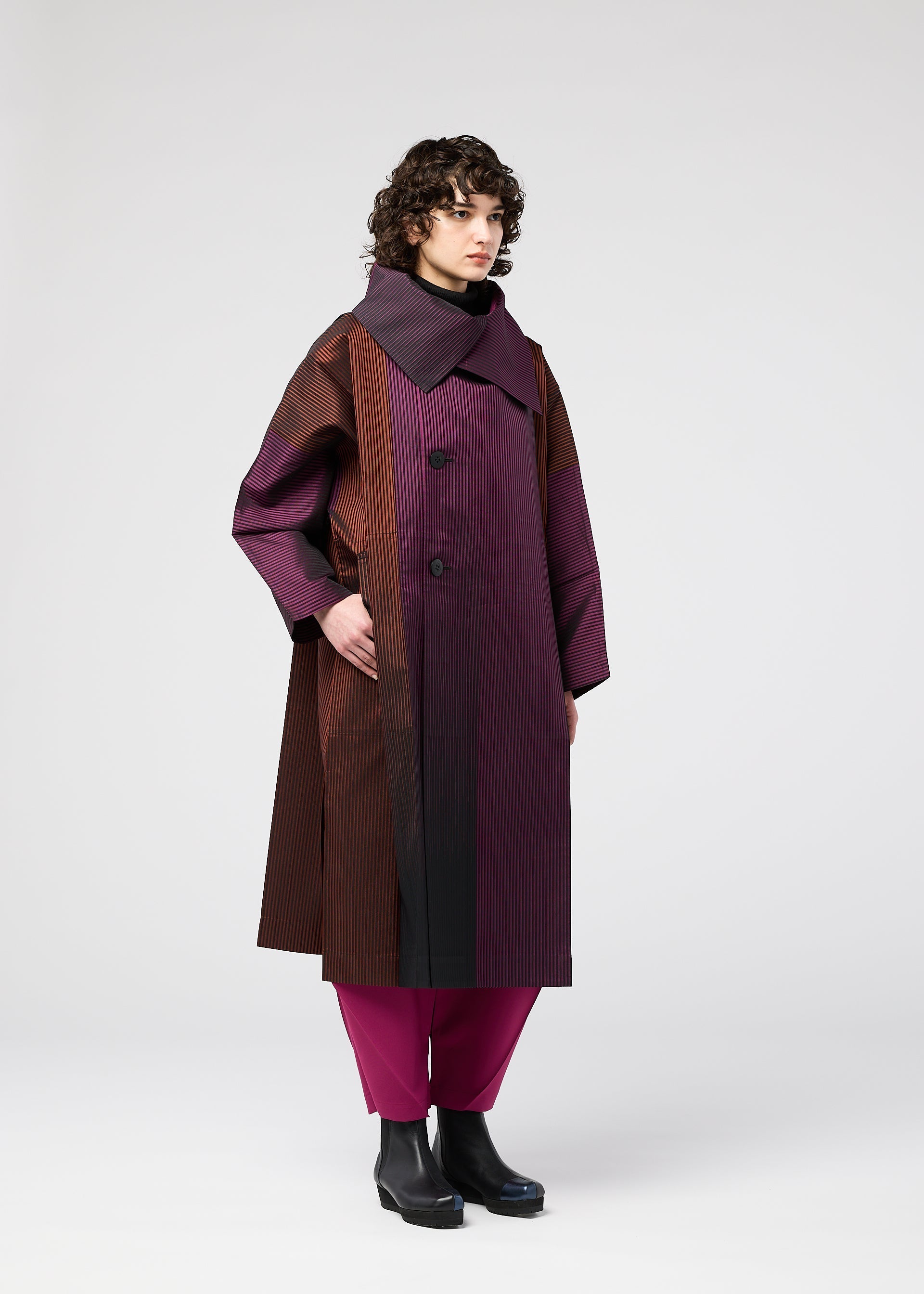 Zoomed view A person with curly hair models the GRADATION COAT by 132 5. ISSEY MIYAKE, a long, oversized jacquard coat with vertical stripes in burgundy, brown, and black. Styled with magenta pants and black boots against a plain white background.