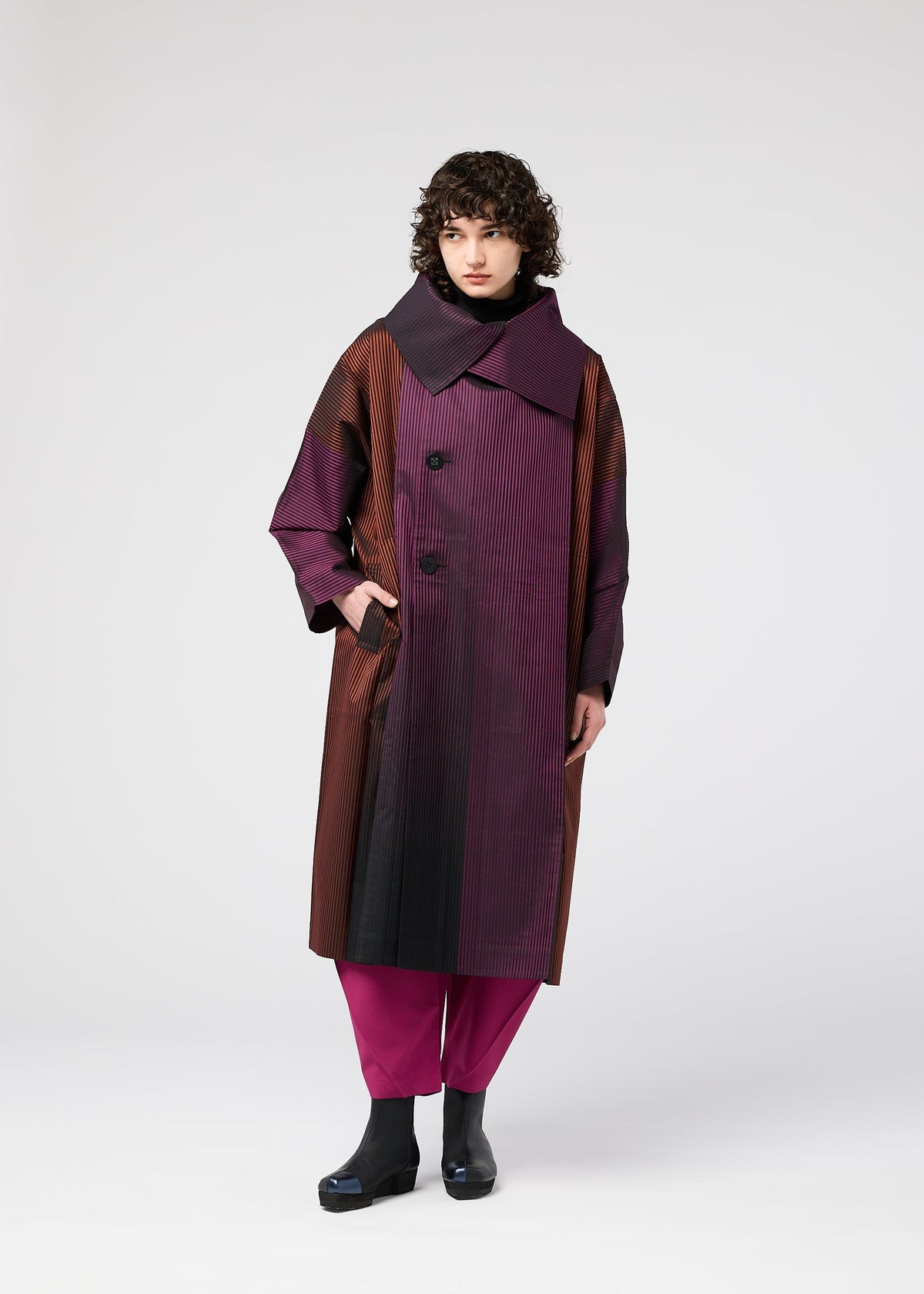 A person with curly hair wears the 132 5. ISSEY MIYAKE GRADATION COAT, long and oversized with a large collar in burgundy, brown, and black hues, styled with magenta pants and black shoes against a plain light background. view 10 of 11