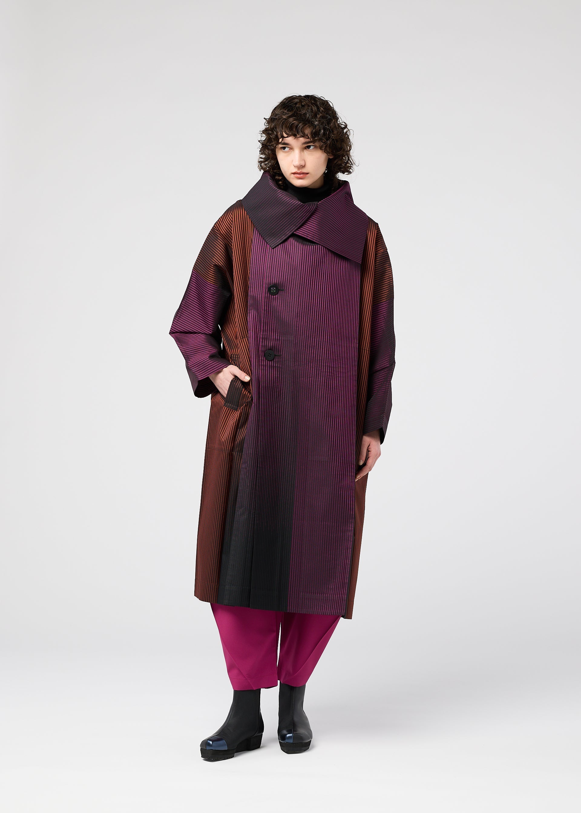 Zoomed view A person with curly hair wears the 132 5. ISSEY MIYAKE GRADATION COAT, long and oversized with a large collar in burgundy, brown, and black hues, styled with magenta pants and black shoes against a plain light background.