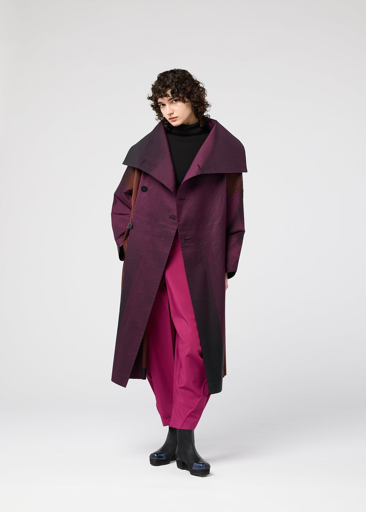 A person with curly dark hair wears the GRADATION COAT by 132 5. ISSEY MIYAKE over magenta pants, a black turtleneck, and black boots, standing against a plain light gray background. view 11 of 11