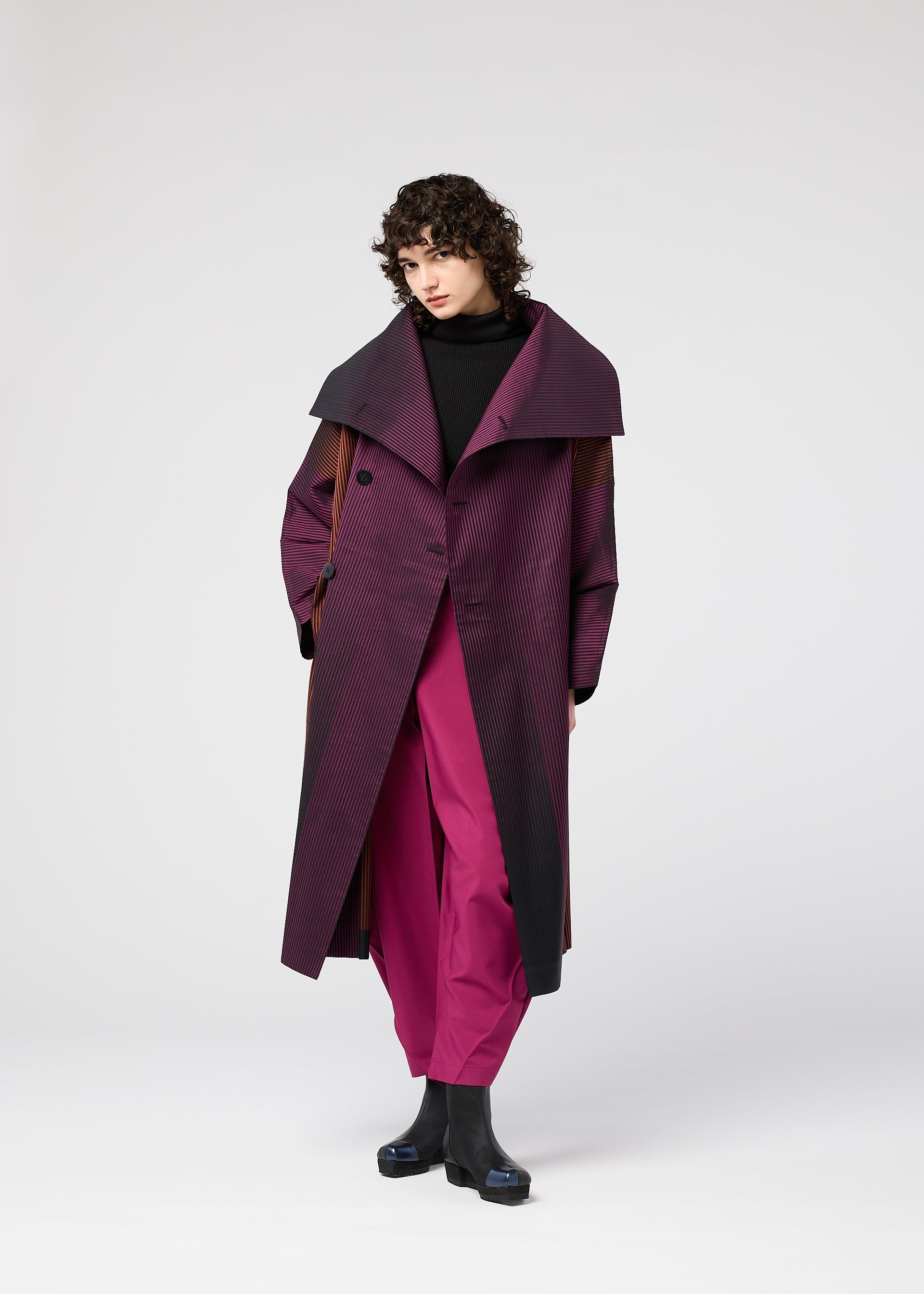 Zoomed view A person with curly dark hair wears the GRADATION COAT by 132 5. ISSEY MIYAKE over magenta pants, a black turtleneck, and black boots, standing against a plain light gray background.