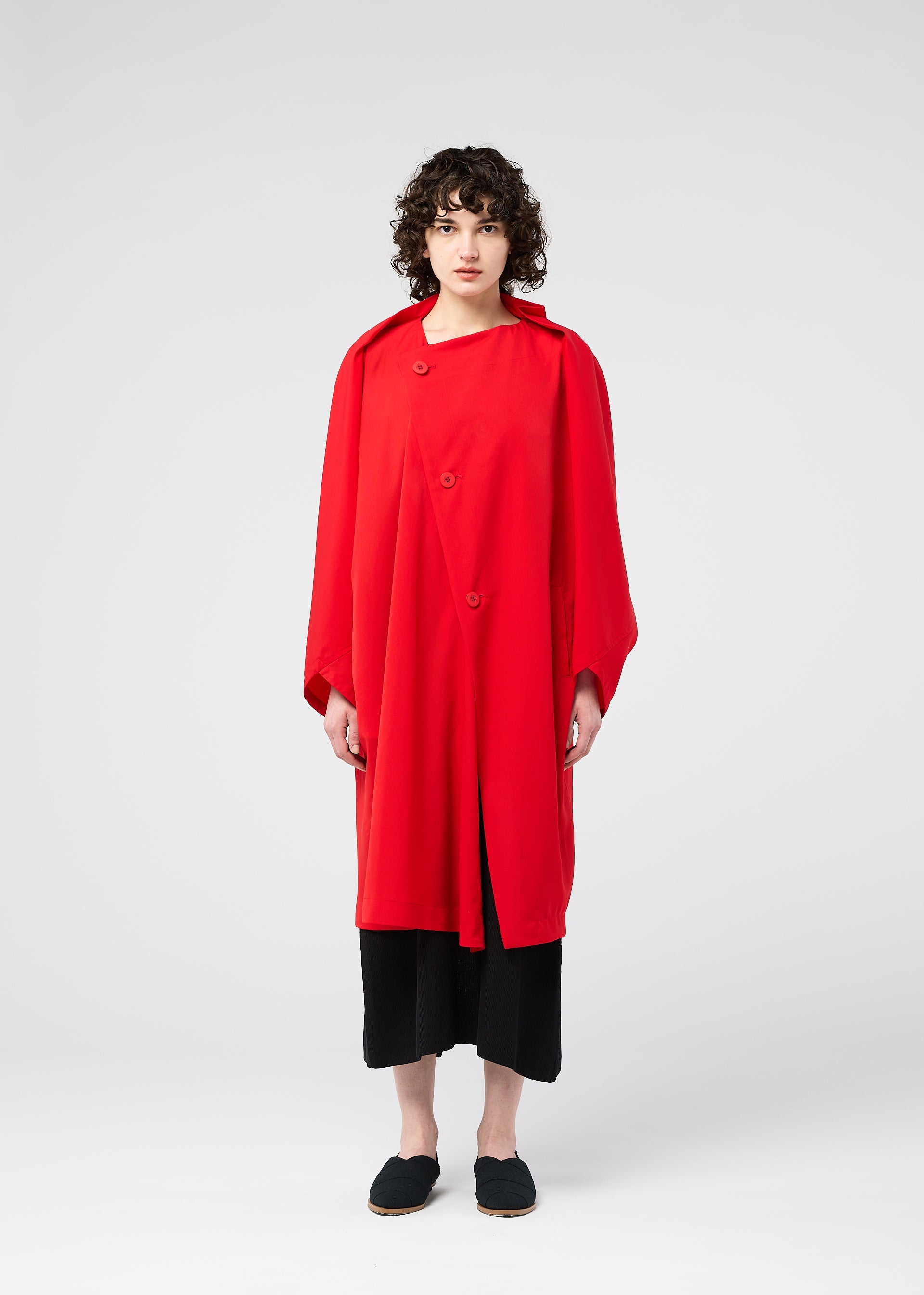 Zoomed view A person with curly hair faces forward, wearing the PARALLEL WRAP by 132 5. ISSEY MIYAKE—a loose-fitting, bright red lightweight coat—over black pants and black slip-on shoes, set against a plain white background.