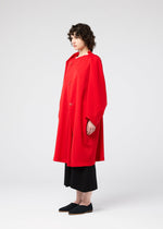A person with short curly hair stands sideways against a plain white background, wearing the PARALLEL WRAP coat by 132 5. ISSEY MIYAKE with black wide-leg pants and black slip-on shoes.